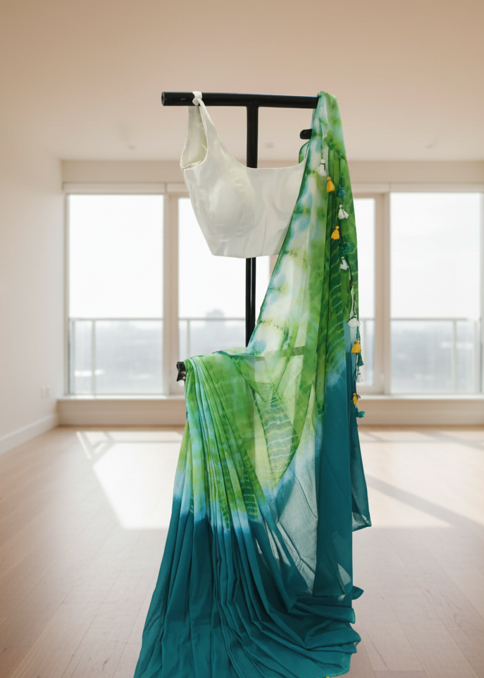 Green and blue gradient mul cotton saree with yellow tassels on a black stand against a white wall.