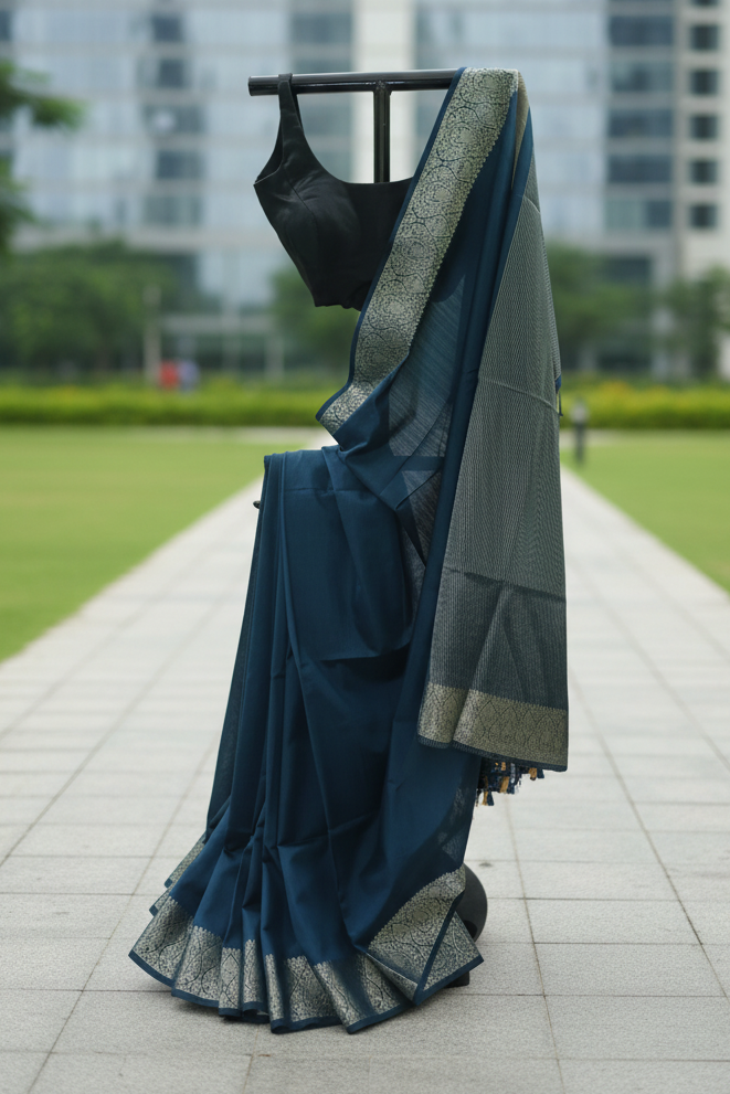 A dark teal blue saree with a black blouse and a silver patterned border draped over a clothing stand, set against a blurred background of a modern glass building and green park grass.