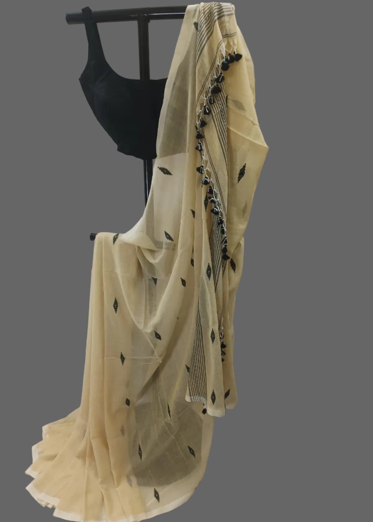 A cream-colored sheer saree with black leaf and border prints, draped over a black dress form against a white curtain backdrop.