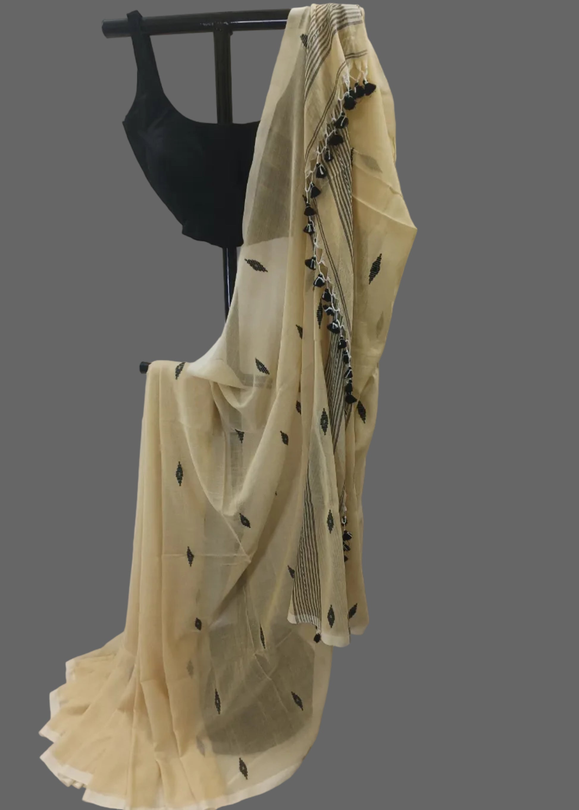 A cream-colored sheer saree with black leaf and border prints, draped over a black dress form against a white curtain backdrop.