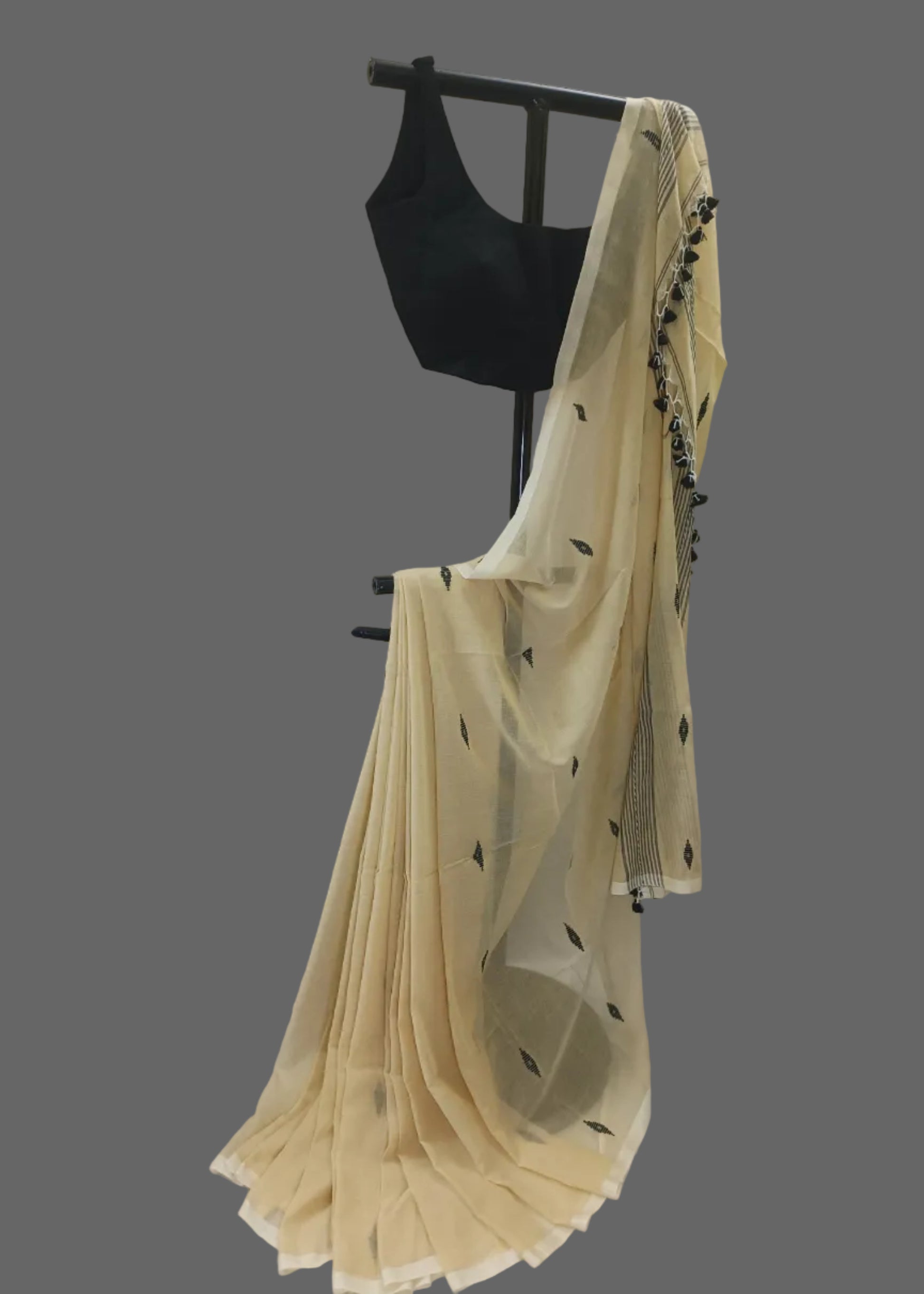 A cream-colored saree with small black leaf motifs and a black embroidered border, draped over a black dress form with a black sleeveless blouse, set against a white draped fabric backdrop.