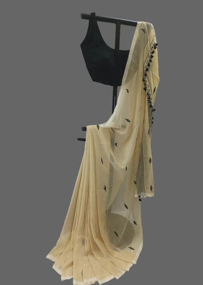 A cream-colored saree with small black leaf motifs and a black embroidered border, draped over a black dress form with a black sleeveless blouse, set against a white draped fabric backdrop.