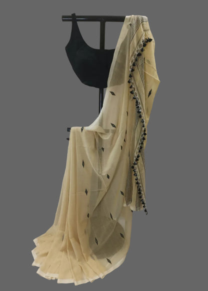 A cream-colored semi-sheer saree with black printed motifs and a black tassel border, draped on a black dress form with a black sleeveless blouse against a draped off-white fabric backdrop.