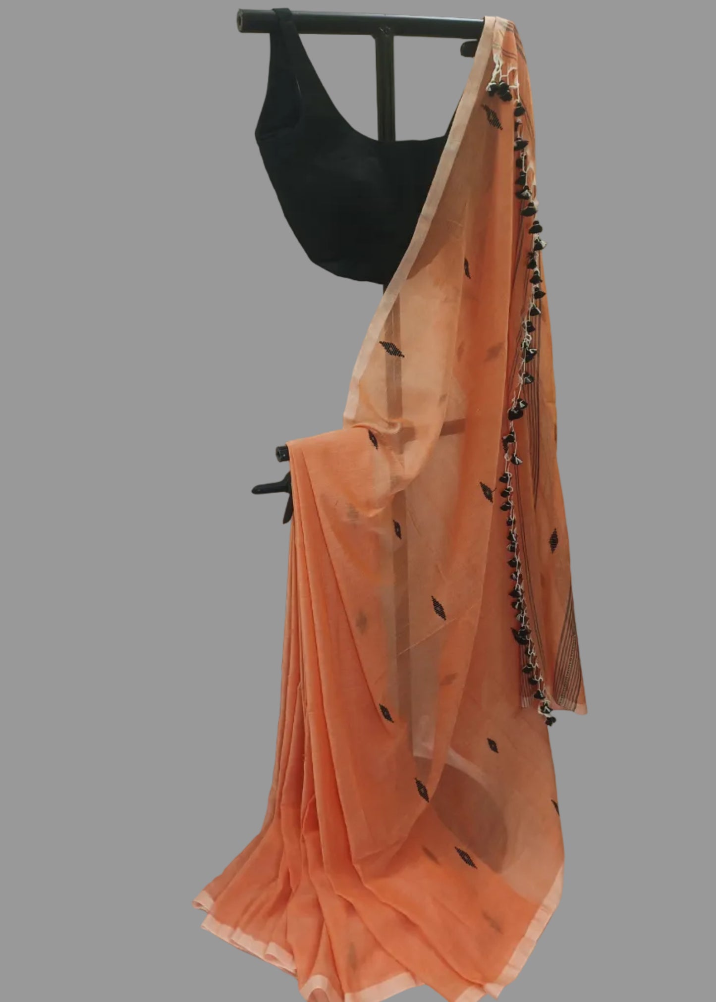 An orange cotton saree with black leaf prints and a black tassel border, draped on a black clothing stand alongside a black sleeveless blouse, set against a white draped fabric backdrop.