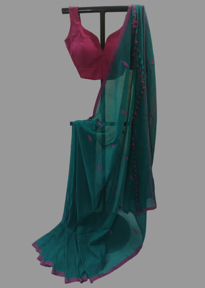 A teal green cotton saree with a magenta pink blouse and border details, displayed on a black clothing rack against a white curtain backdrop.