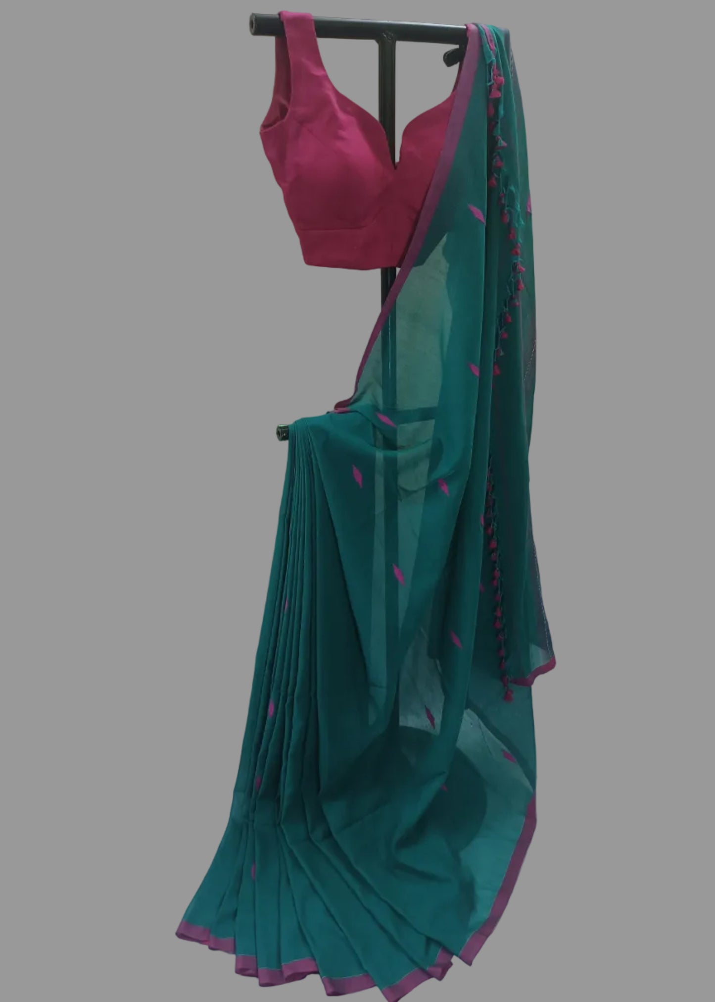 A teal green saree with a magenta pink blouse and border, displayed on a black clothing rack against a white curtain backdrop.