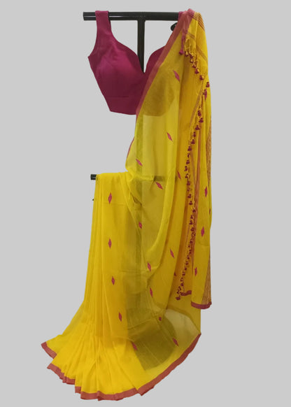 A yellow saree with pink border trim and small pink motifs, paired with a magenta blouse, displayed on a black hanger against a white draped fabric backdrop.
