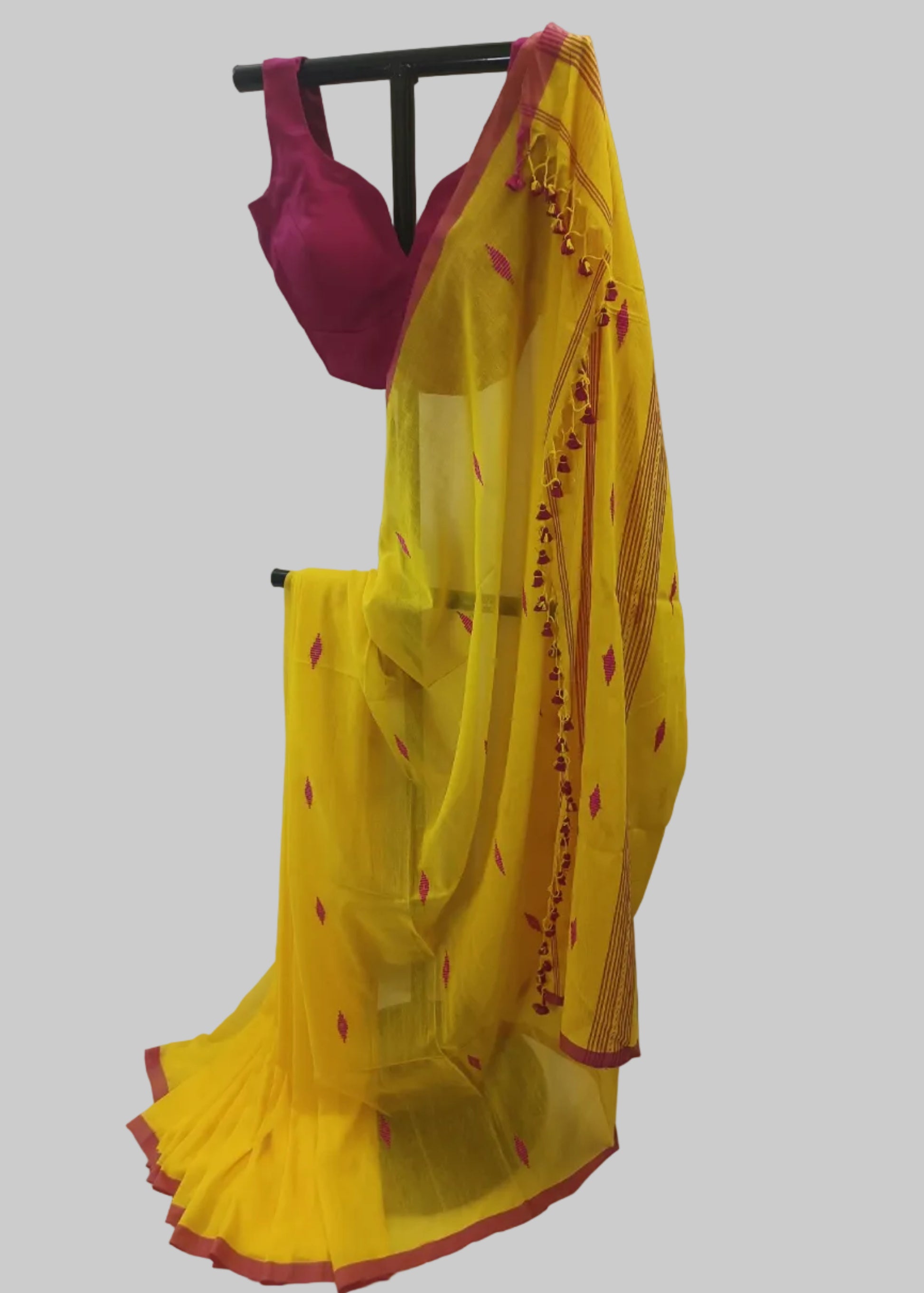 A yellow cotton saree with red border trim and small red motifs, paired with a magenta pink blouse, displayed on a black clothing rack against a white curtain backdrop.