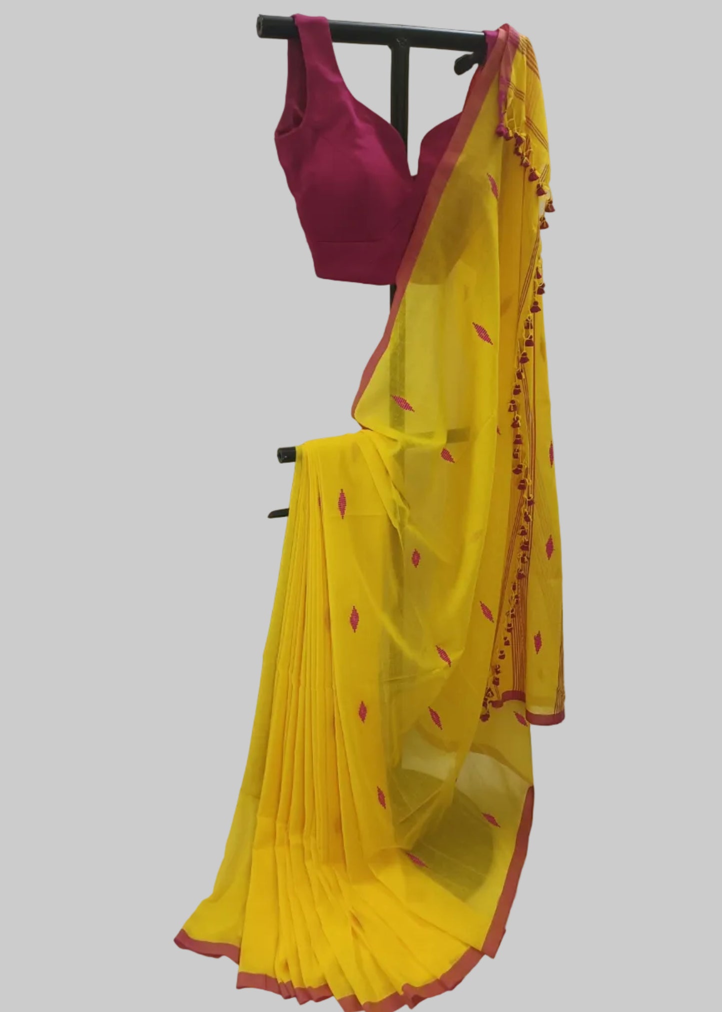 A yellow saree with red border and small red motifs, paired with a magenta pink blouse, displayed on a black clothing rack against a white fabric backdrop.