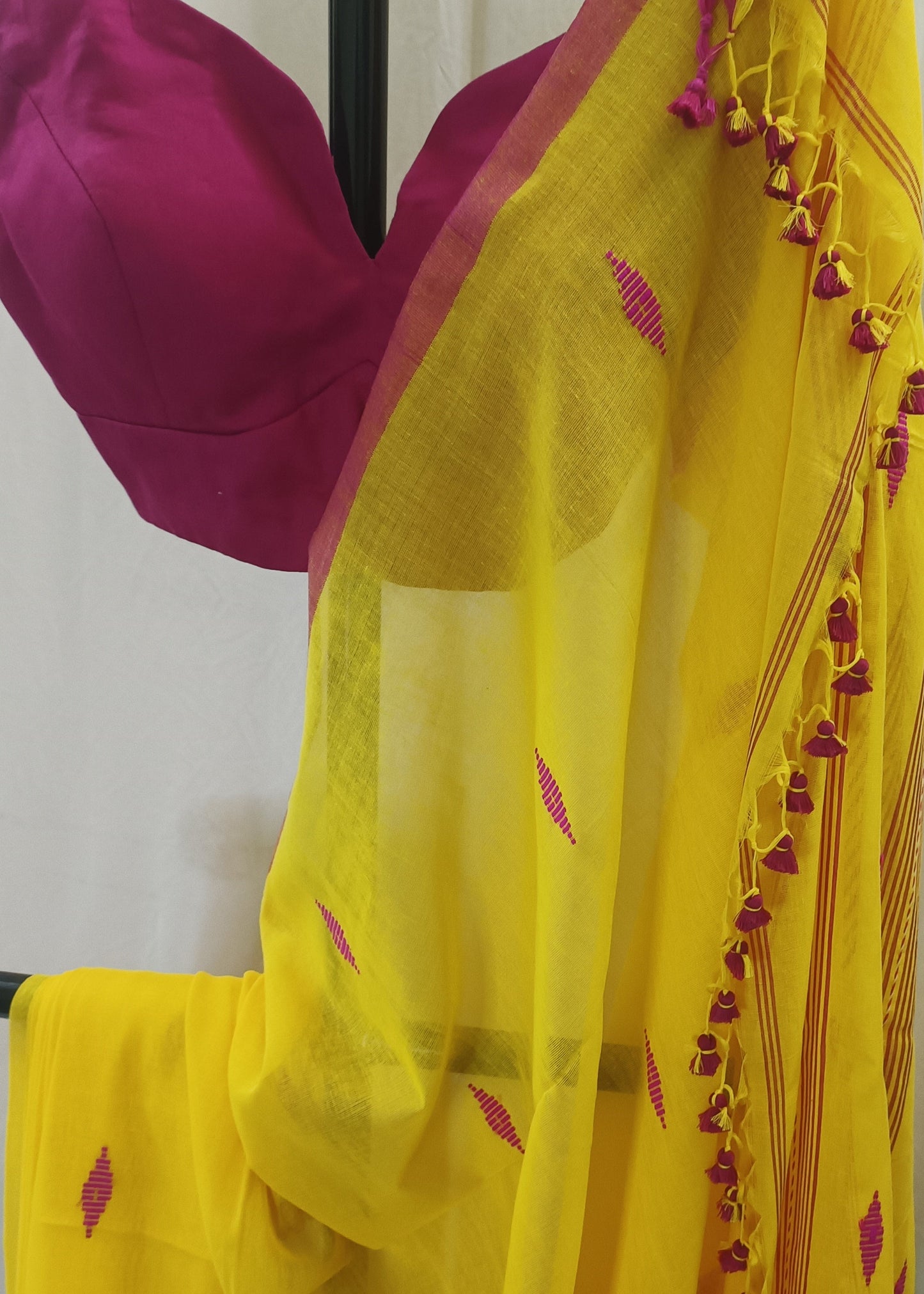 Yellow saree with pink border and small pink diamond motifs, draped over a black hanger with a pink blouse visible at the top, against a light curtain background.