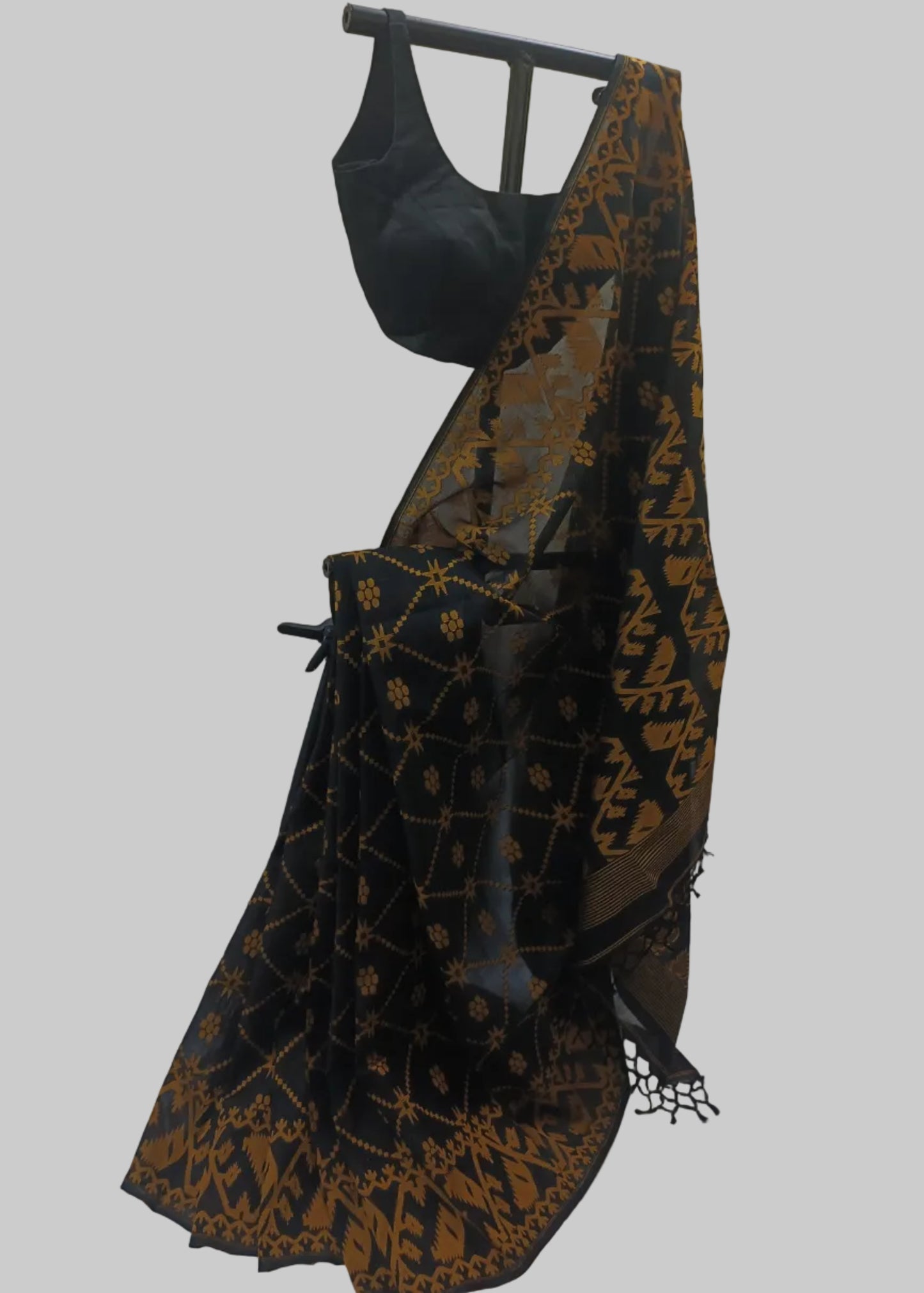 A black saree with a gold geometric and floral pattern, draped on a black hanger stand with a matching black blouse, set against a beige curtain backdrop.