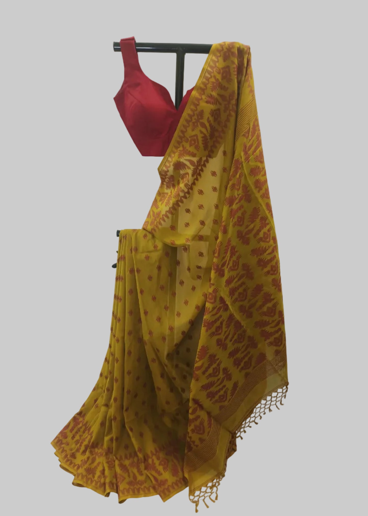 A mustard yellow and red patterned saree draped on a black hanger with a red blouse, set against a white curtain backdrop.