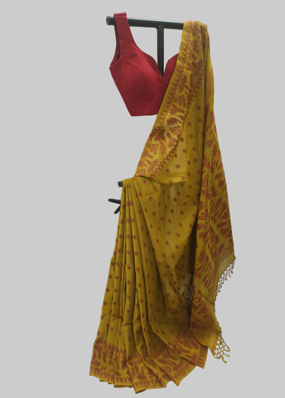 A mustard yellow and brown patterned saree draped on a black clothing stand, paired with a red sleeveless blouse, set against a white fabric backdrop.