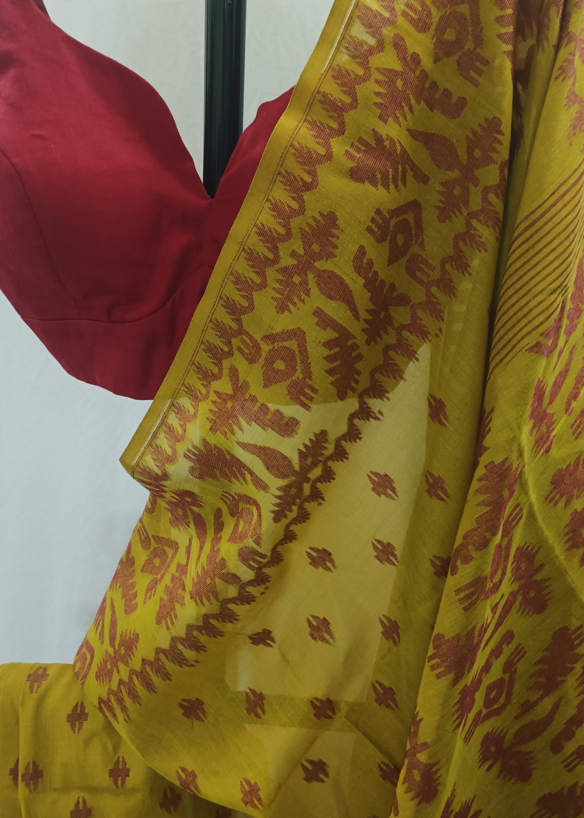 A yellow saree with a rust-red ikat pattern and border, draped over a black hanger with a red blouse visible at the top left, set against a plain off-white background.