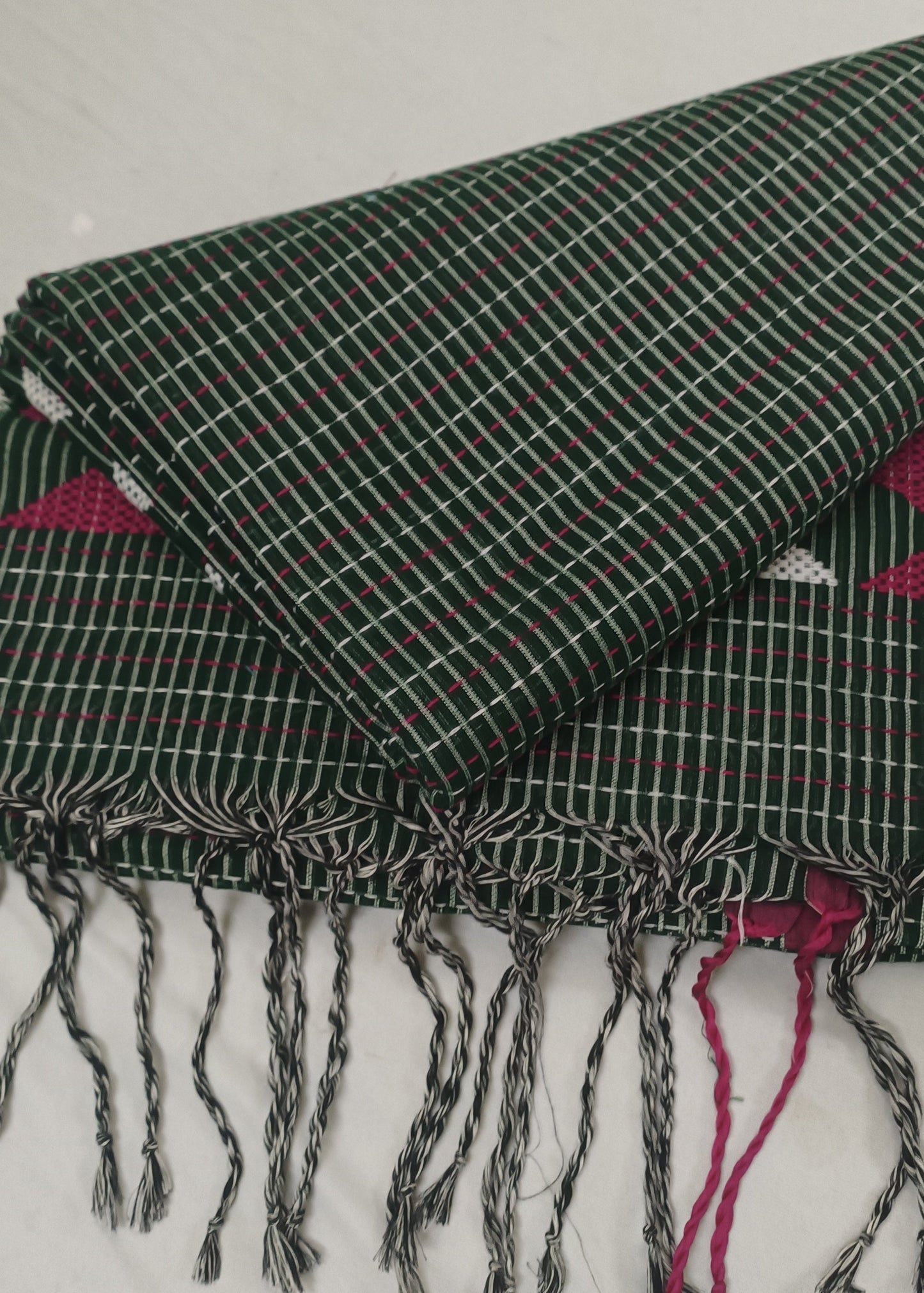 A folded dark green woven textile with white grid lines and fringed tassels, laid on a white fabric background. Pink patterned fabric is visible underneath at the edges.