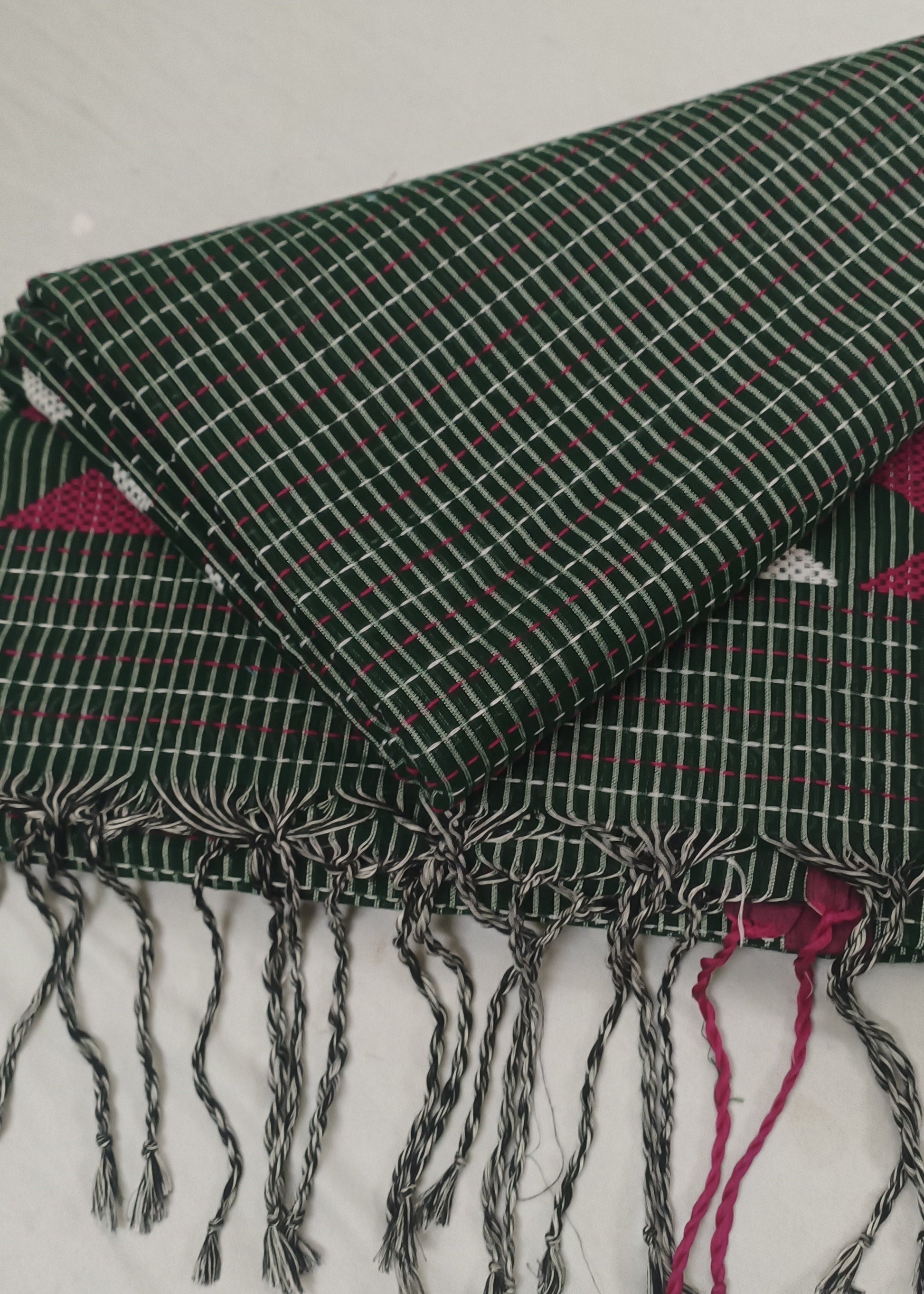 A folded dark green woven textile with white grid lines and fringed tassels, laid on a white fabric background. Pink patterned fabric is visible underneath at the edges.