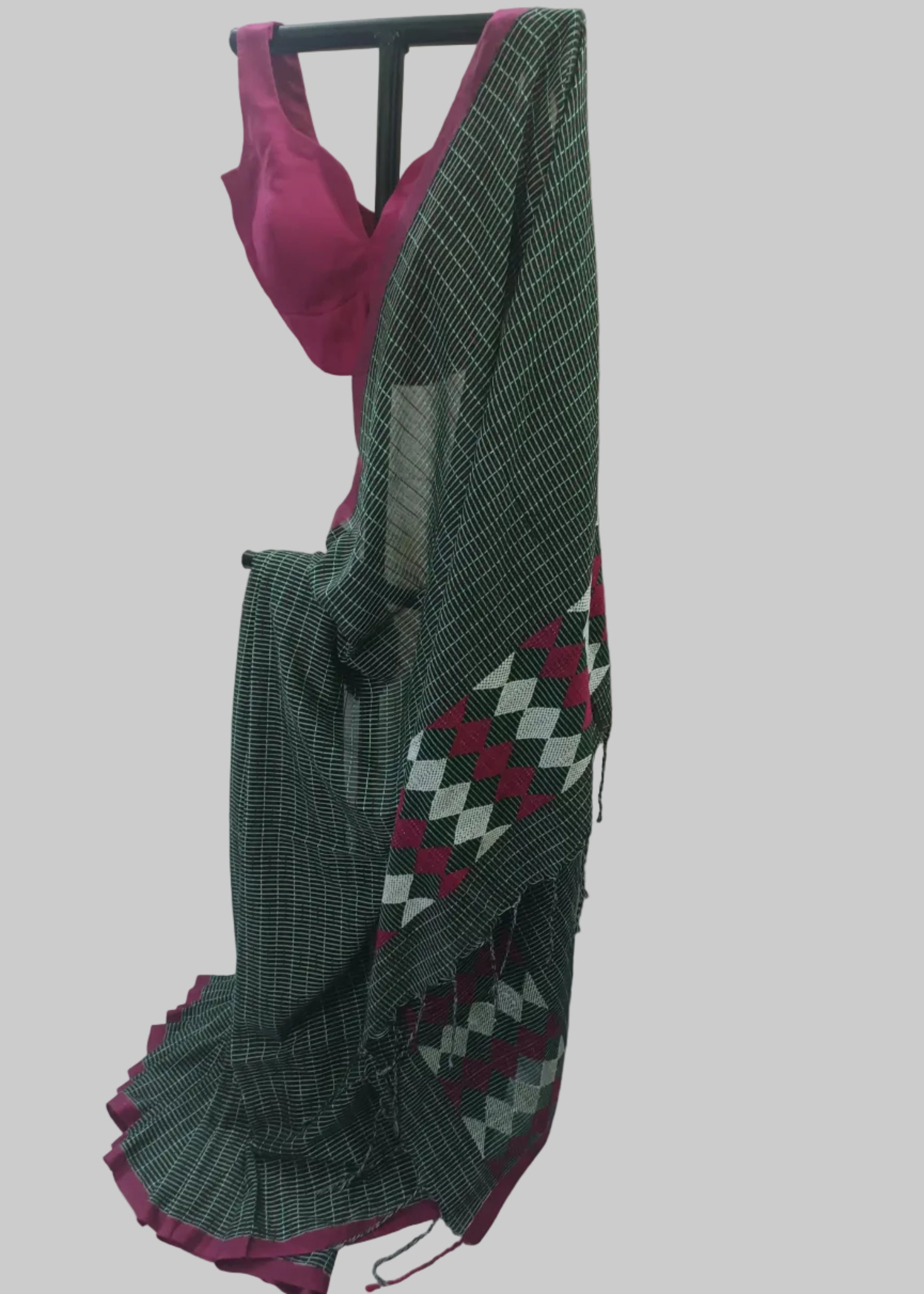 A dark green saree with a white grid pattern and a magenta-pink border, draped on a black hanger with a matching pink blouse visible at the top, set against a draped cream fabric backdrop.