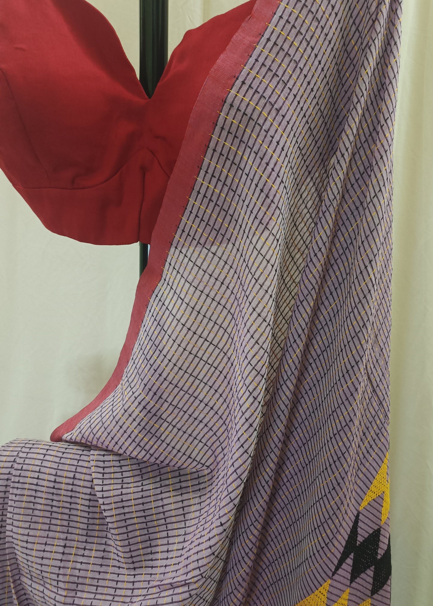 A close-up of a draped saree or fabric panel with a fine purple and white grid pattern, a red border, and a yellow geometric motif at the corner, shown next to a red blouse on a hanger against a cream curtain background.