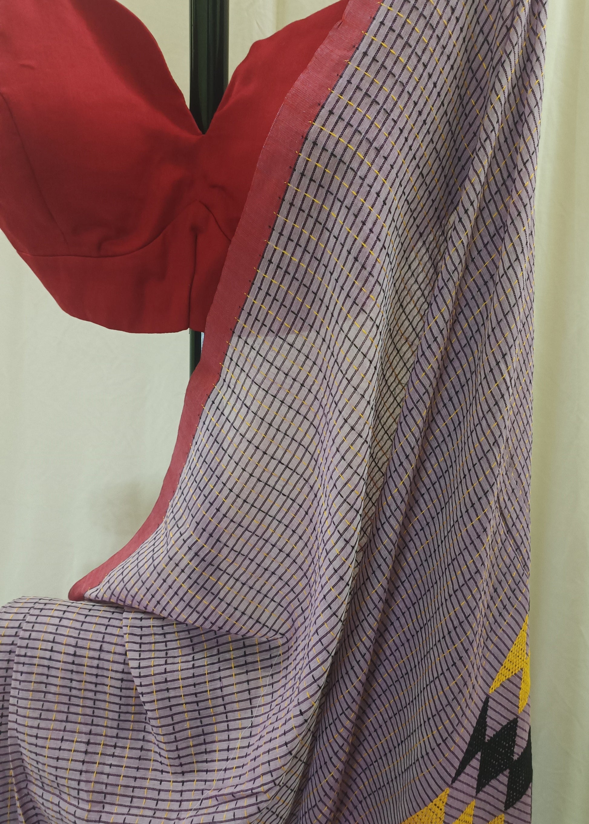 A close-up of a draped saree or fabric panel with a fine purple and white grid pattern, a red border, and a yellow geometric motif at the corner, shown next to a red blouse on a hanger against a cream curtain background.