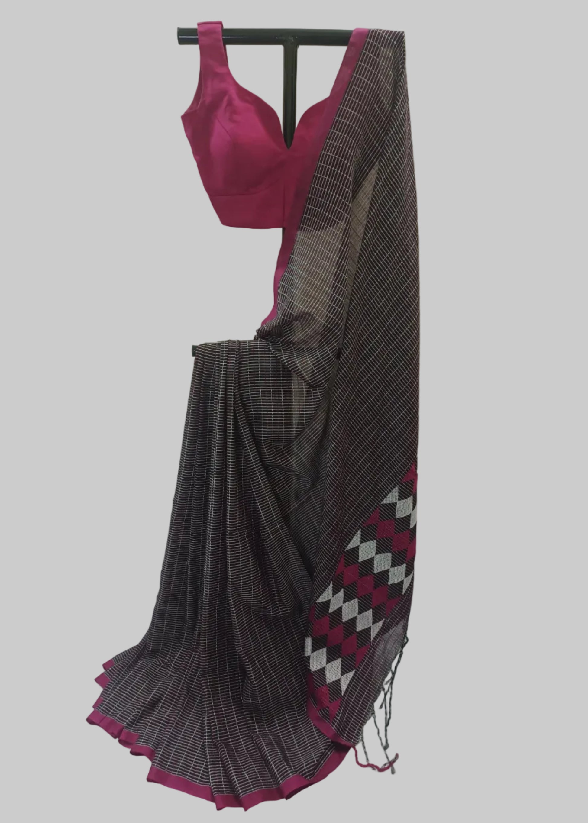 A dark gray and black checkered saree with a magenta pink border and matching blouse, displayed on a black hanger against a cream-colored curtain backdrop.