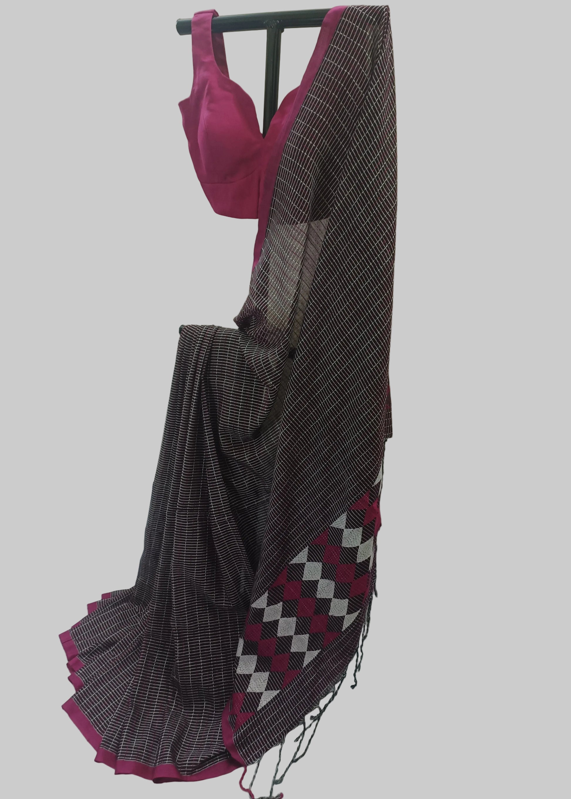 A dark gray and white patterned saree draped on a black hanger with a bright pink blouse, set against a white curtain backdrop.