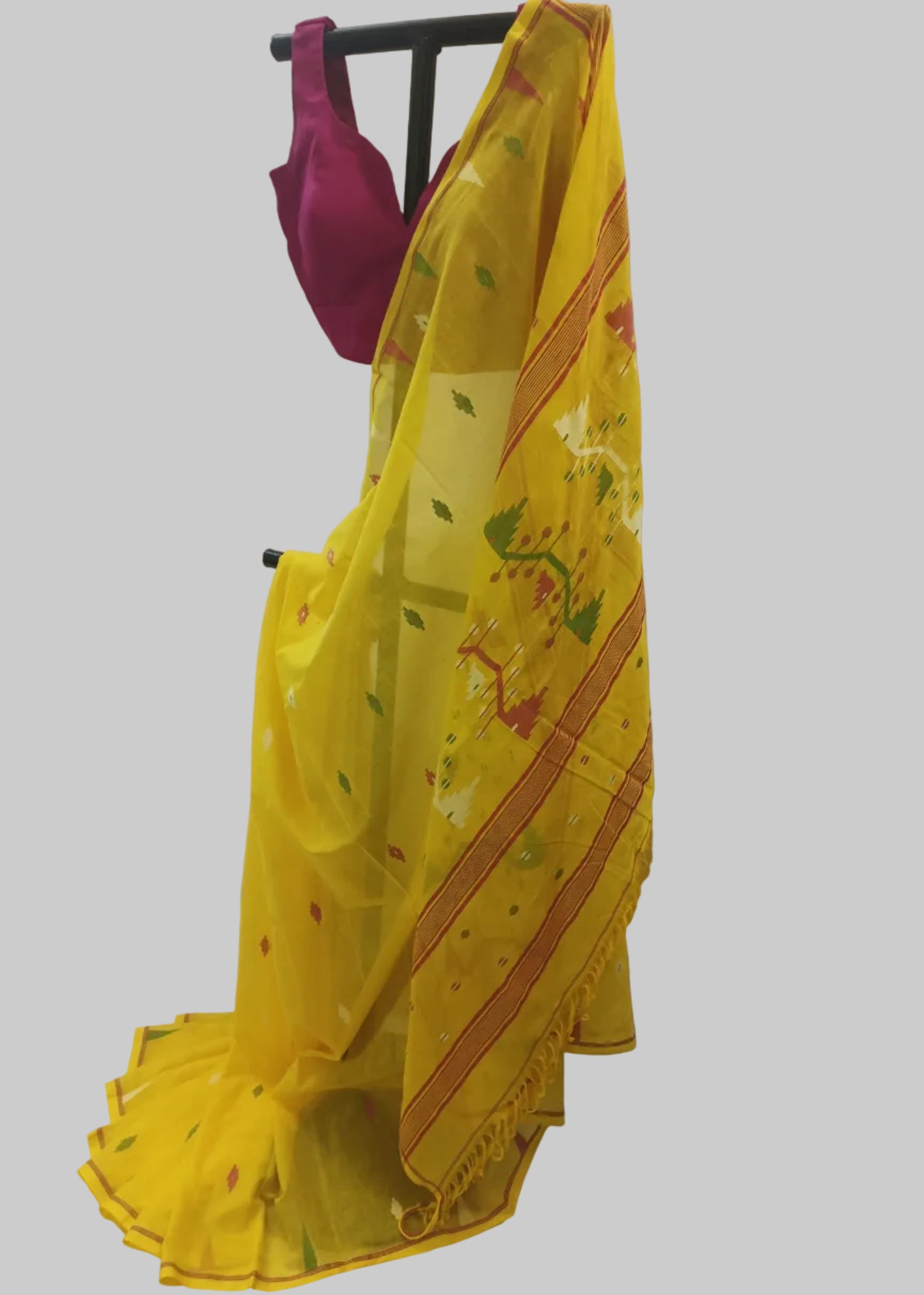 A yellow saree with green and red floral motifs and striped borders, draped on a black clothing stand with a magenta blouse visible at the top, set against a white fabric backdrop.