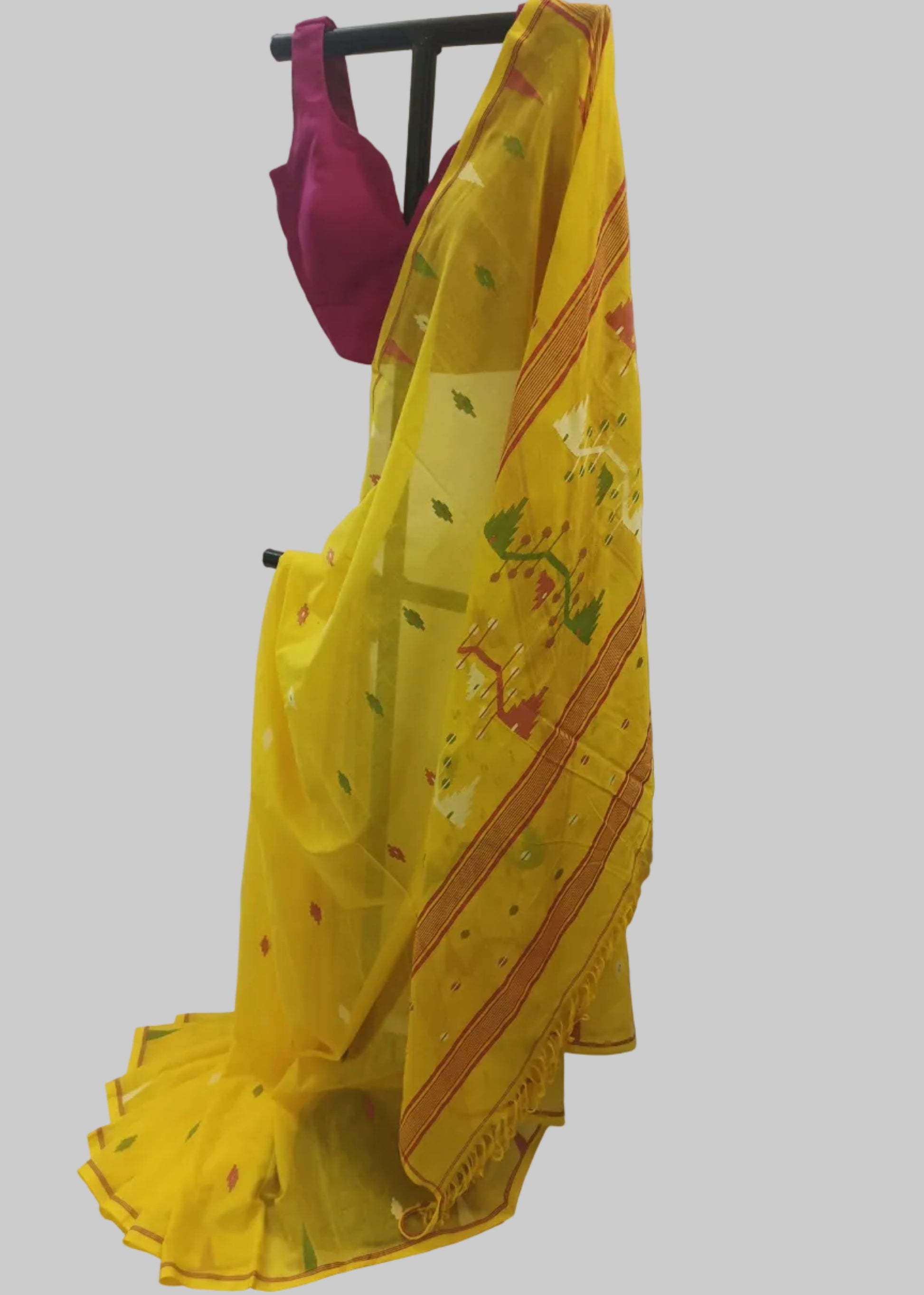 A yellow saree with green and red floral motifs and striped borders, draped on a black clothing stand with a magenta blouse visible at the top, set against a white fabric backdrop.