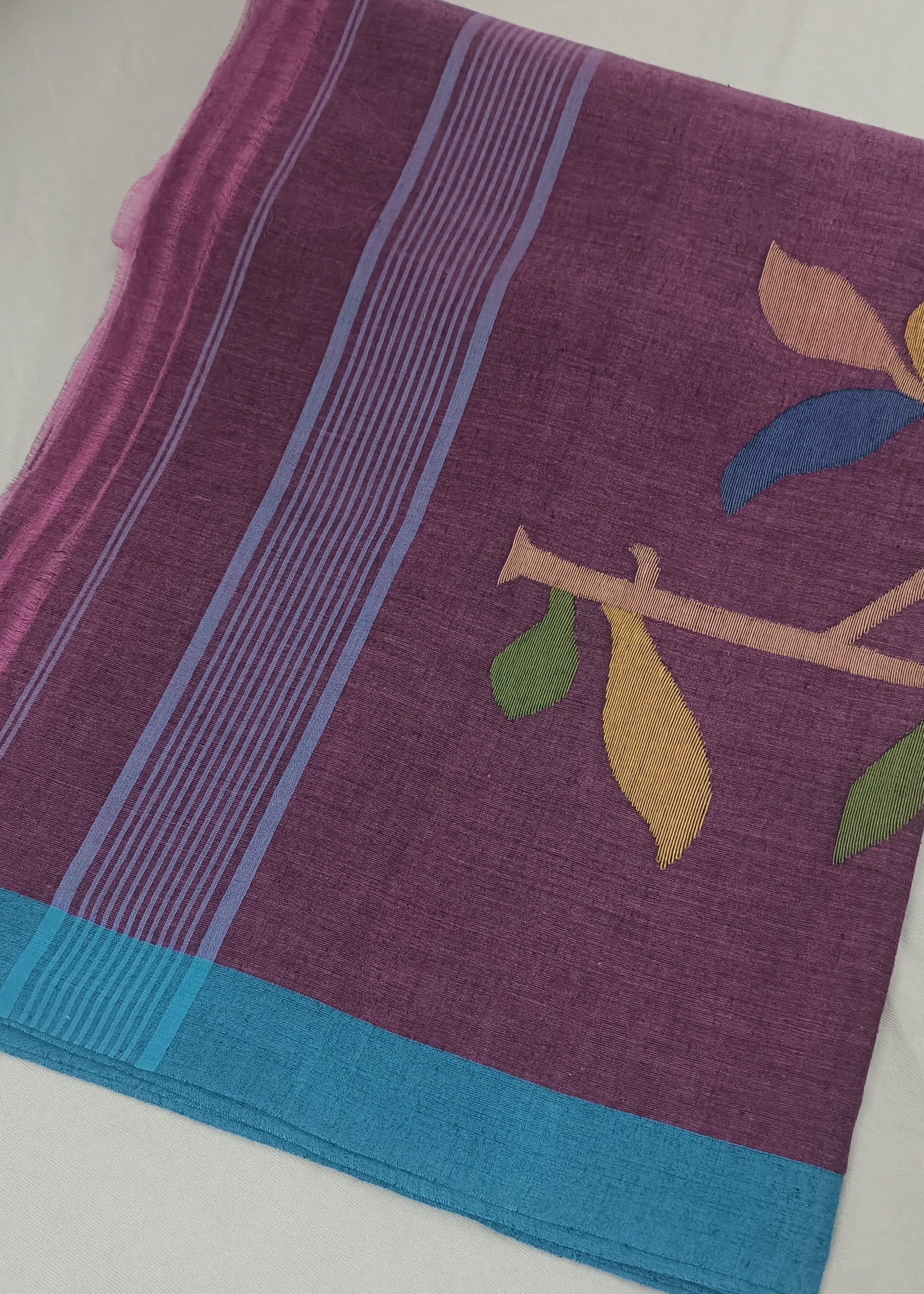 A folded purple saree or fabric cloth with a turquoise blue border, light blue stripes, and a printed branch motif with yellow, green, and blue leaves, laid flat on a white surface.