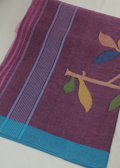 A folded purple saree or fabric cloth with a turquoise blue border, light blue stripes, and a printed branch motif with yellow, green, and blue leaves, laid flat on a white surface.