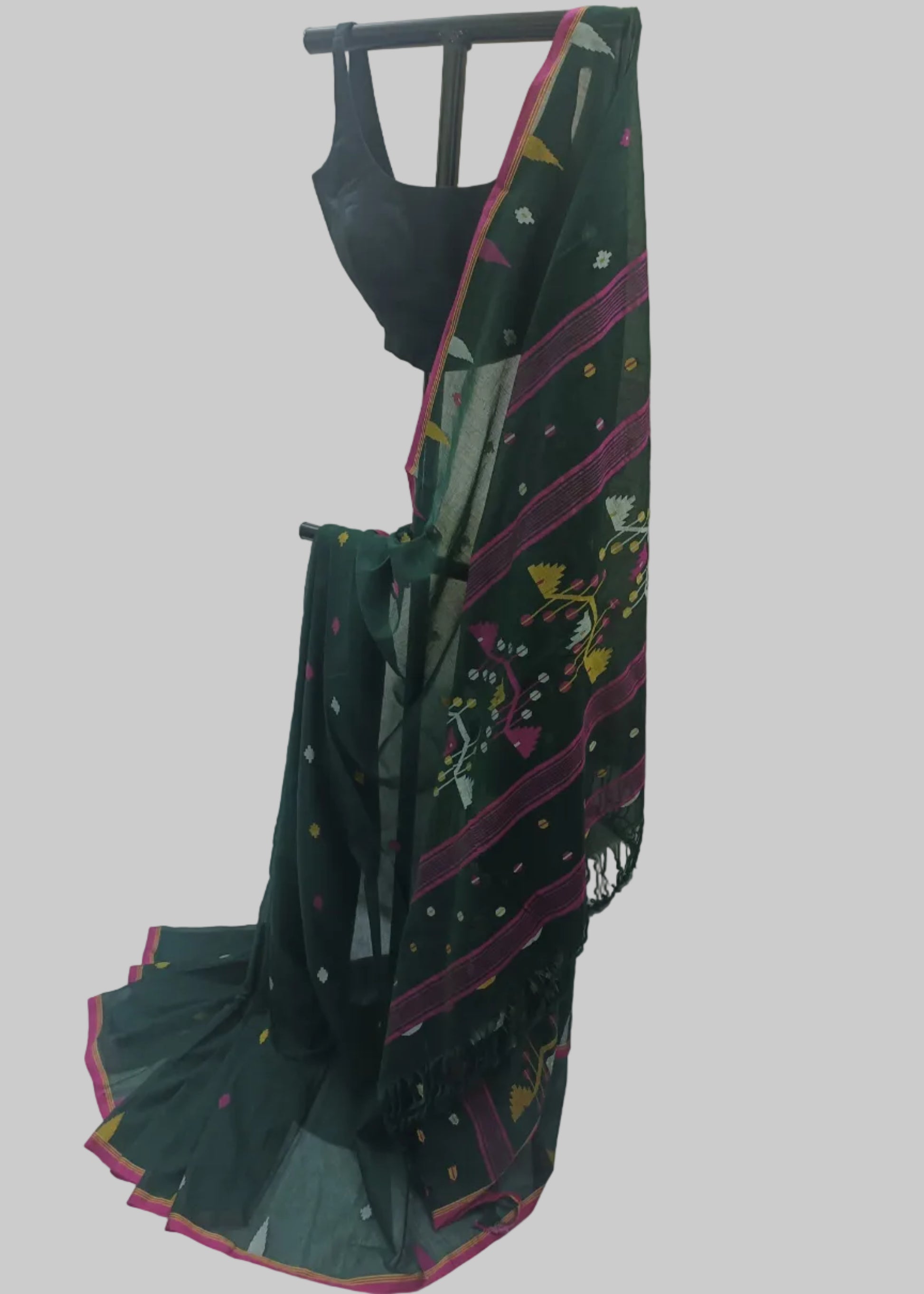 A dark green saree with pink border stripes, multicolor floral prints, and small dotted motifs, draped on a black dress form with a black blouse. The backdrop is a white fabric sheet.