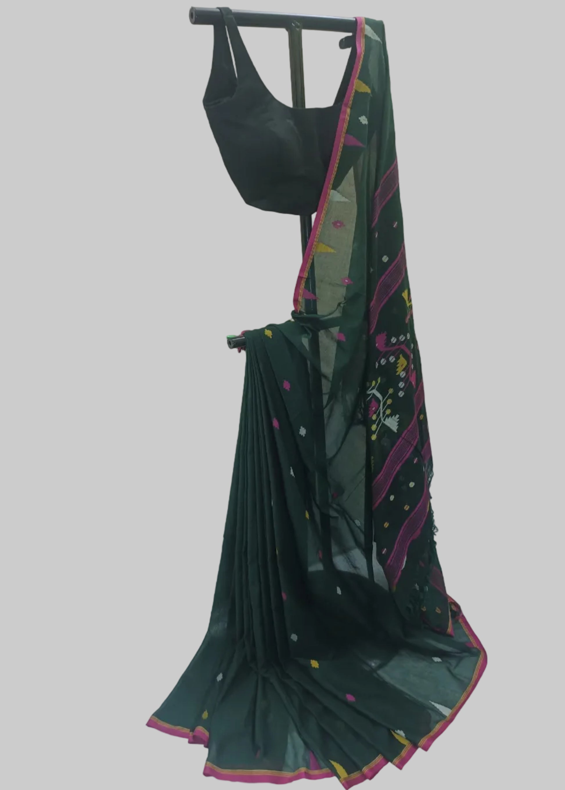 A dark green saree with pink and multicolor border patterns, paired with a black sleeveless blouse, displayed on a black clothing rack against a draped white fabric backdrop.
