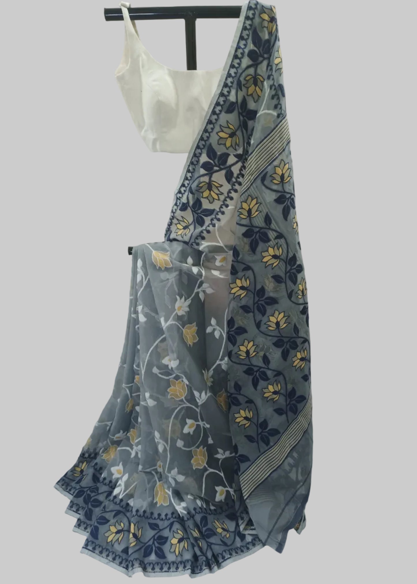 A sheer gray saree with navy blue and yellow floral patterns, draped over a white blouse on a black hanger stand against a white fabric backdrop.