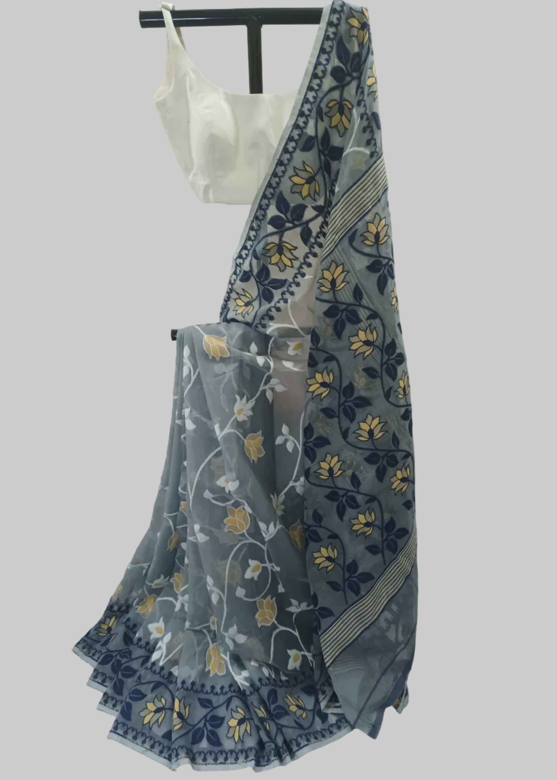 A sheer gray saree with navy blue and yellow floral patterns, draped over a white blouse on a black hanger stand against a white fabric backdrop.