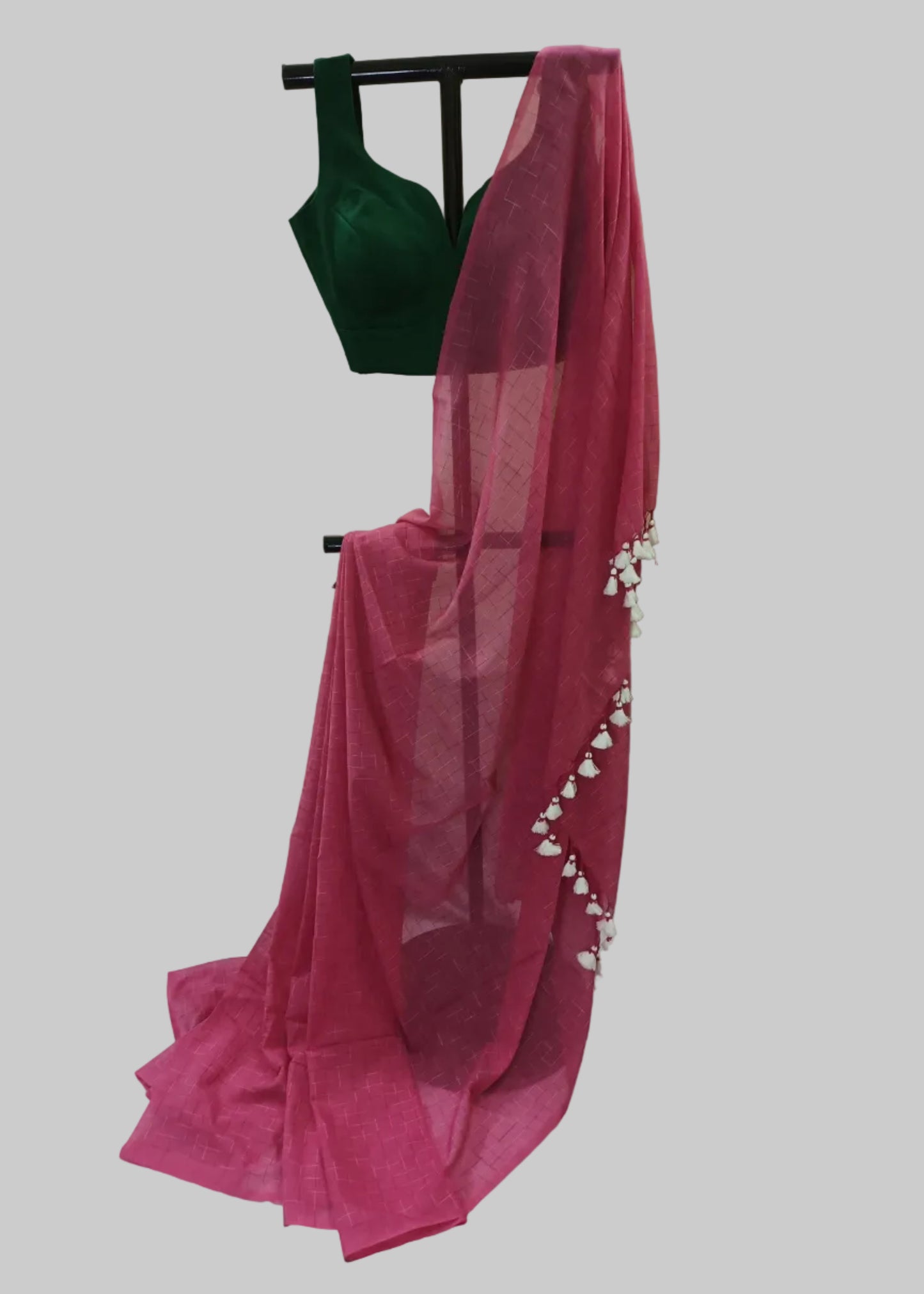 A bright pink sheer dupatta draped over a black hanger, paired with a dark green sleeveless blouse, set against a draped white fabric backdrop.