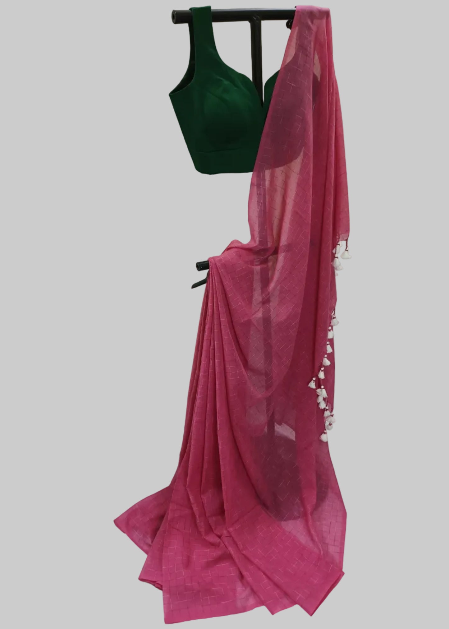 A bright pink sheer saree drape with white floral tassels, paired with a dark green sleeveless blouse, displayed on a black hanger against a draped white fabric backdrop.