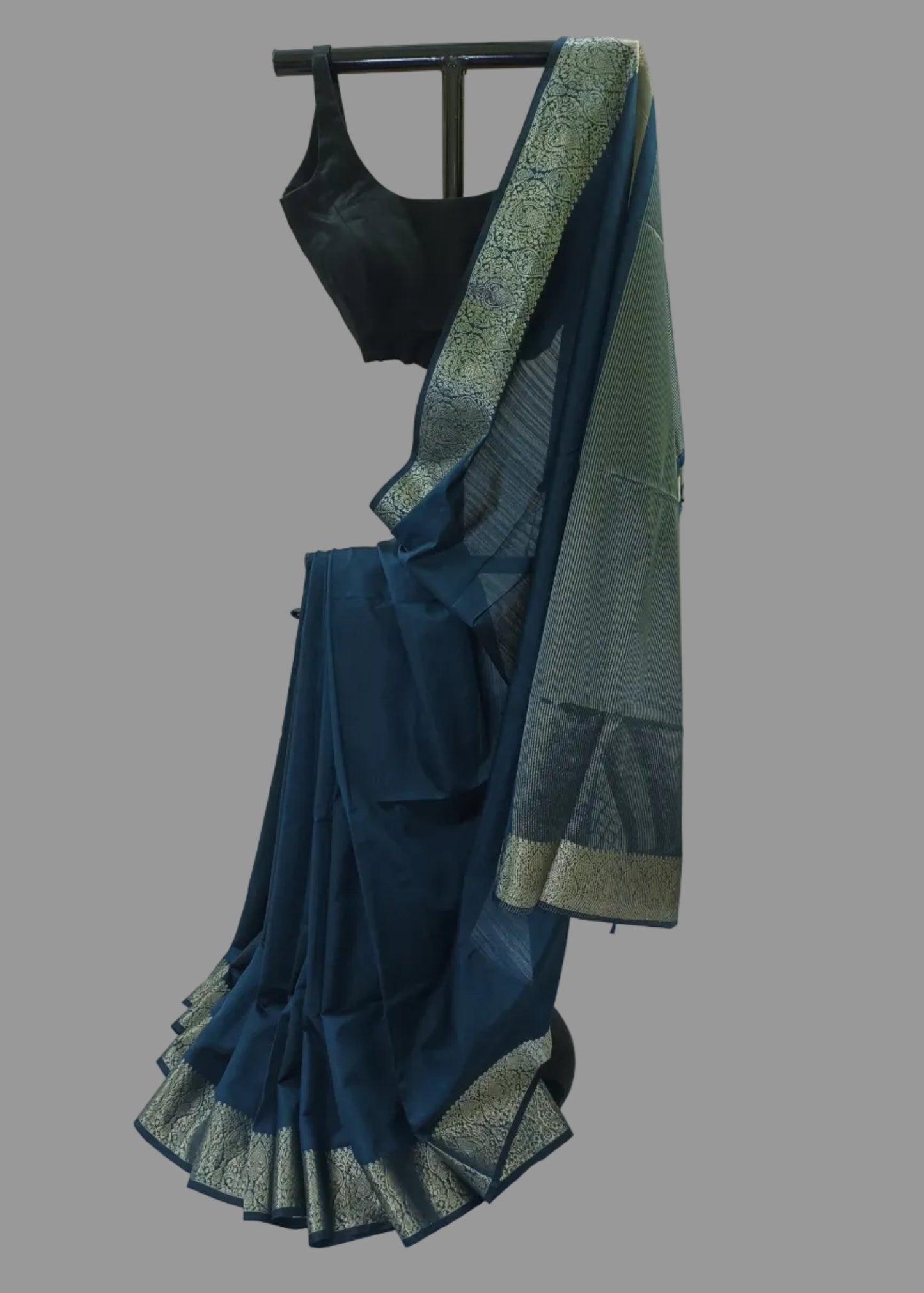 A dark teal blue saree with a wide metallic gold and silver patterned border, paired with a black sleeveless blouse, displayed on a black hanger against a draped white fabric backdrop.