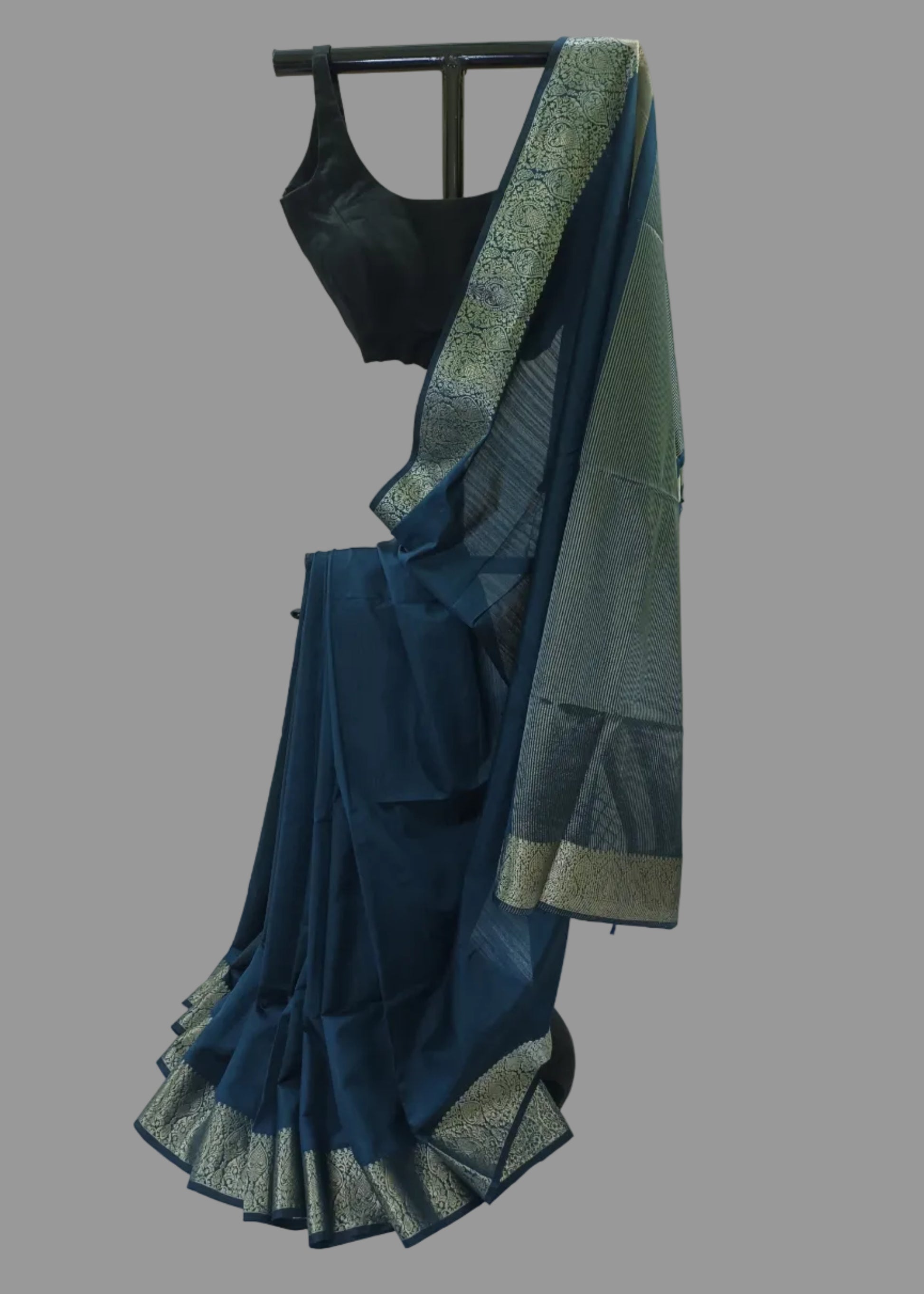 A dark teal blue saree with a wide metallic gold and silver patterned border, paired with a black sleeveless blouse, displayed on a black hanger against a draped white fabric backdrop.