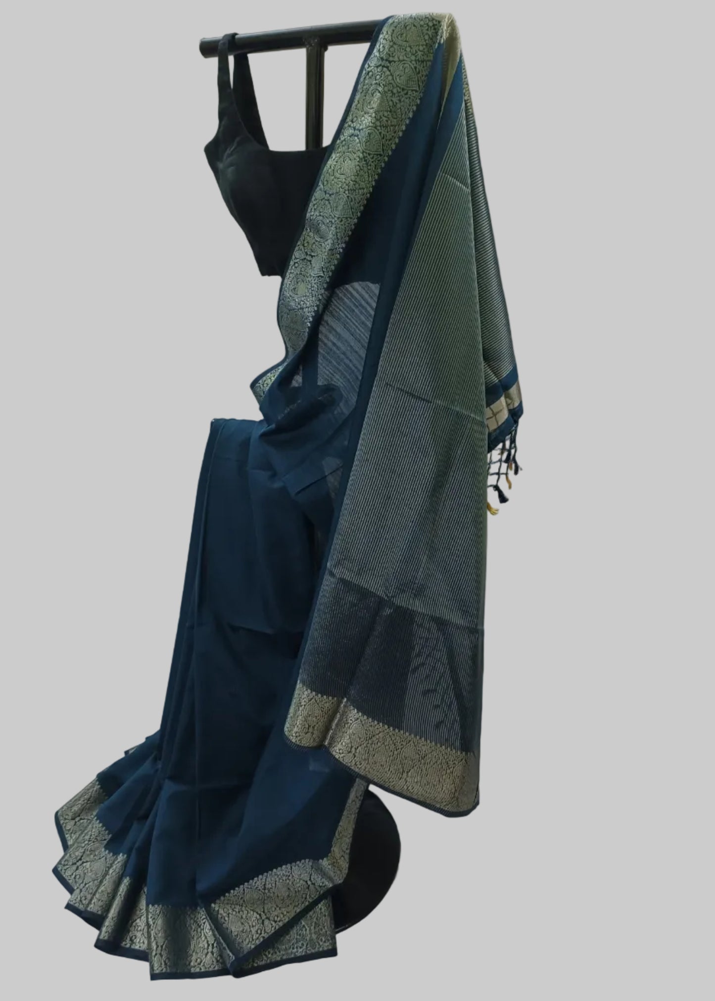 A dark teal blue saree with a wide metallic gold border and a matching teal blouse draped on a black dress form against a white fabric backdrop.