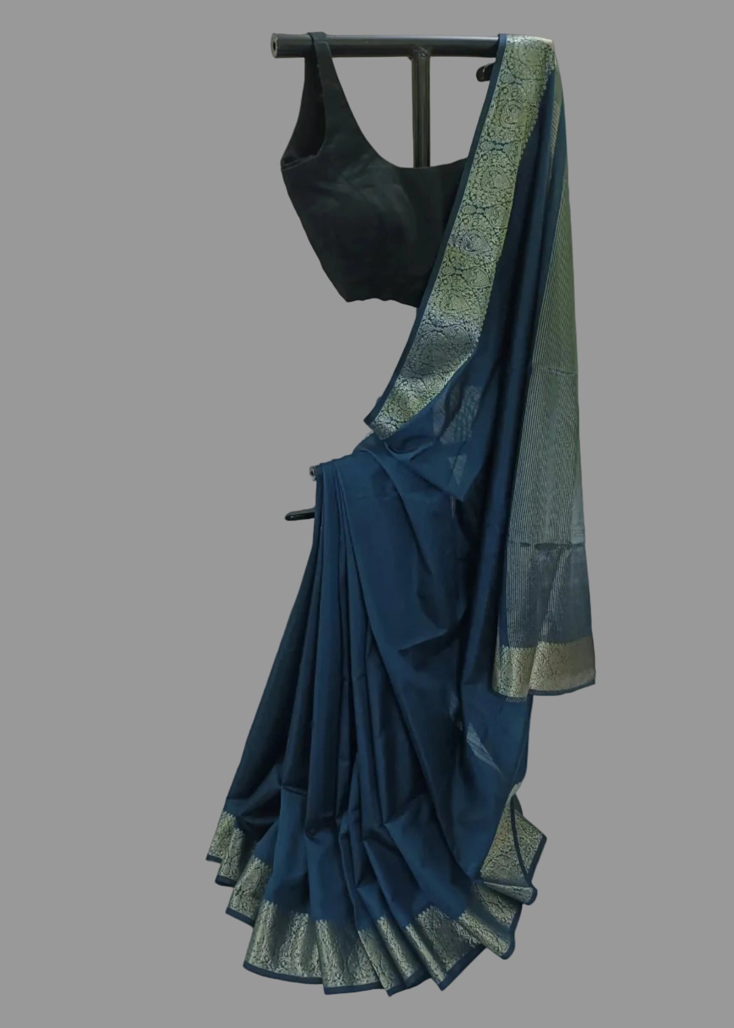 A dark teal blue saree with a wide metallic gold border and a black sleeveless blouse, displayed on a black clothing rack against a draped white fabric backdrop.