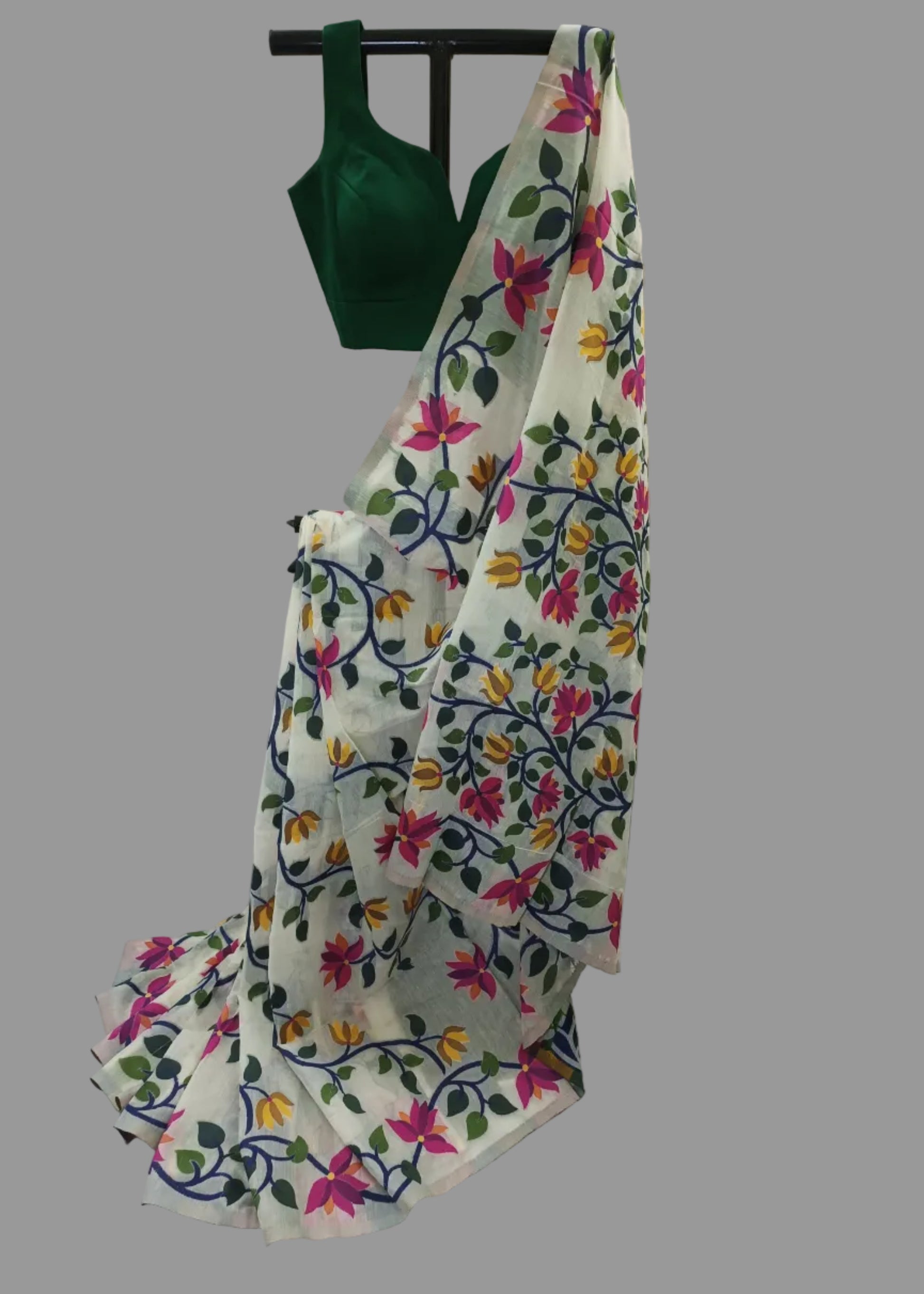 A white saree with colorful floral and vine patterns, draped over a black hanger with a dark green blouse visible at the top, set against a white draped fabric backdrop.