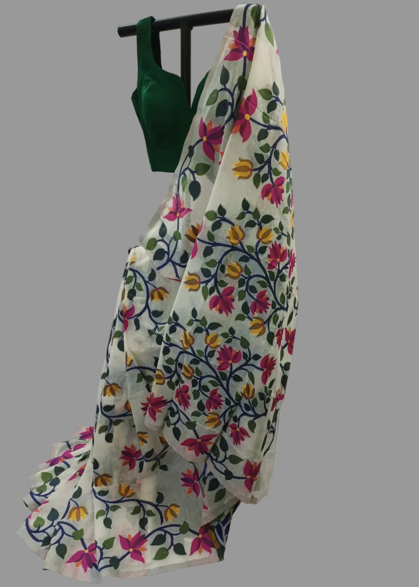A white saree draped over a black hanger, featuring colorful floral embroidery in pink, yellow, green, and blue, with a solid dark green blouse piece hanging at the top against a draped white fabric backdrop.