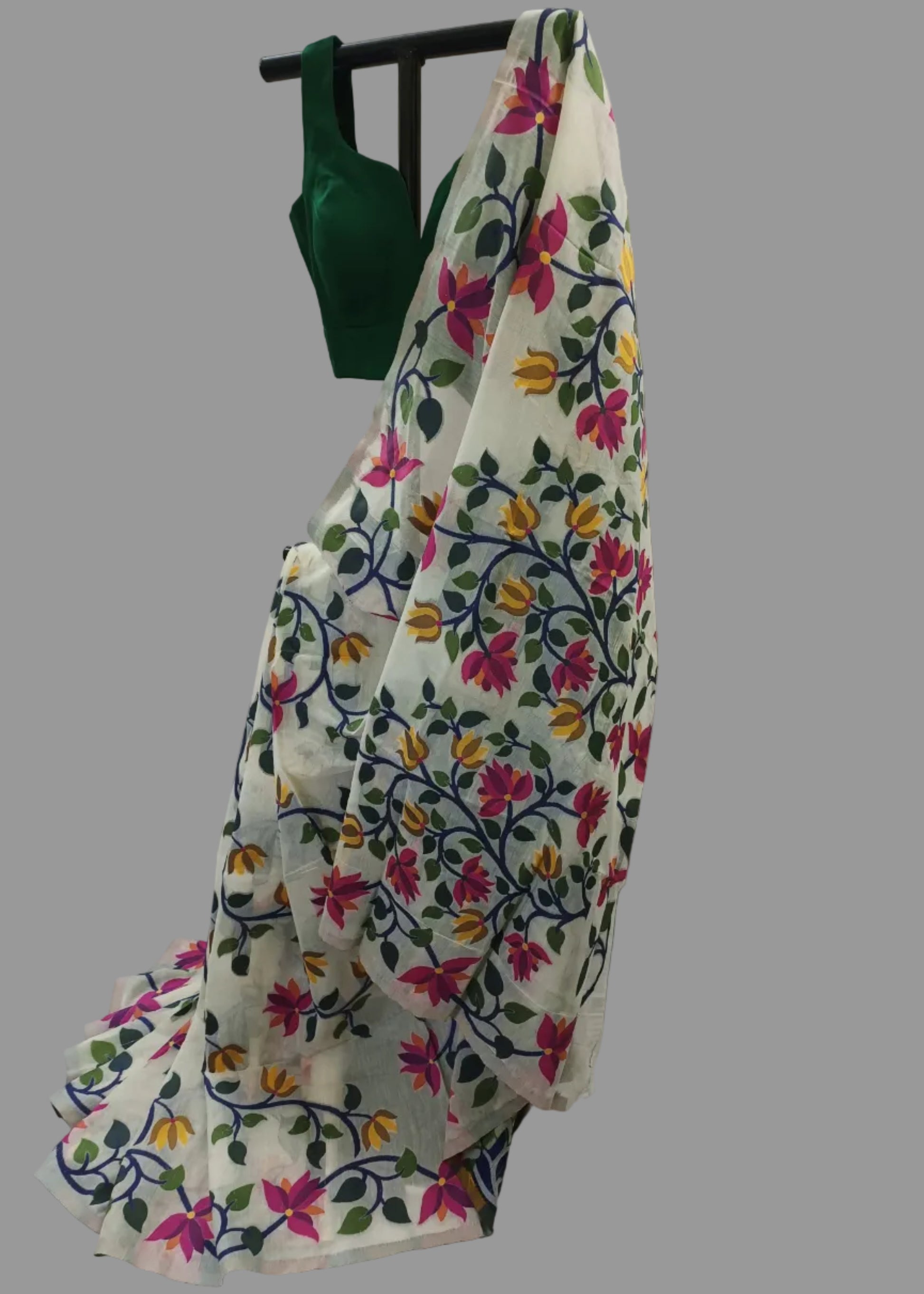 A white saree draped over a black hanger, featuring colorful floral embroidery in pink, yellow, green, and blue, with a solid dark green blouse piece hanging at the top against a draped white fabric backdrop.