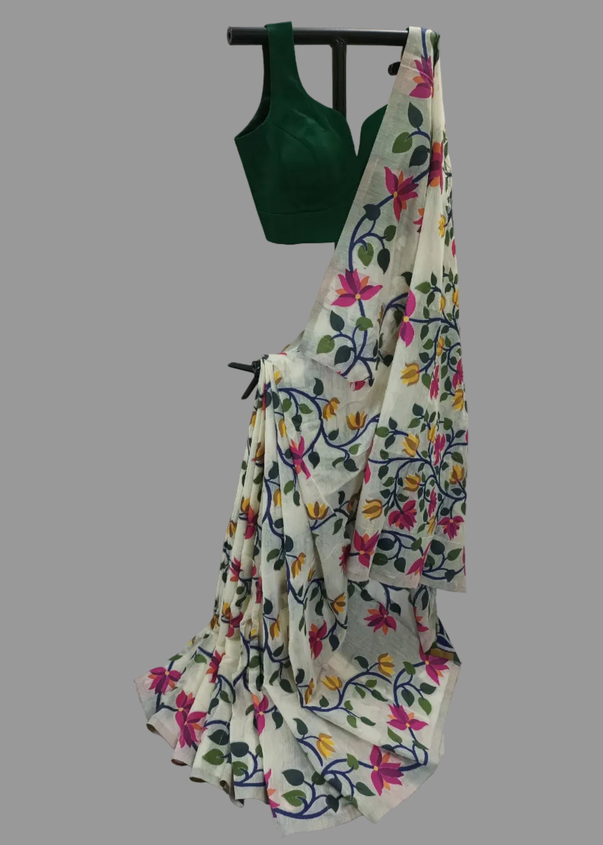 A white saree with a vibrant multicolor floral print, paired with a dark green sleeveless blouse, displayed on a black hanger against a white draped fabric backdrop.