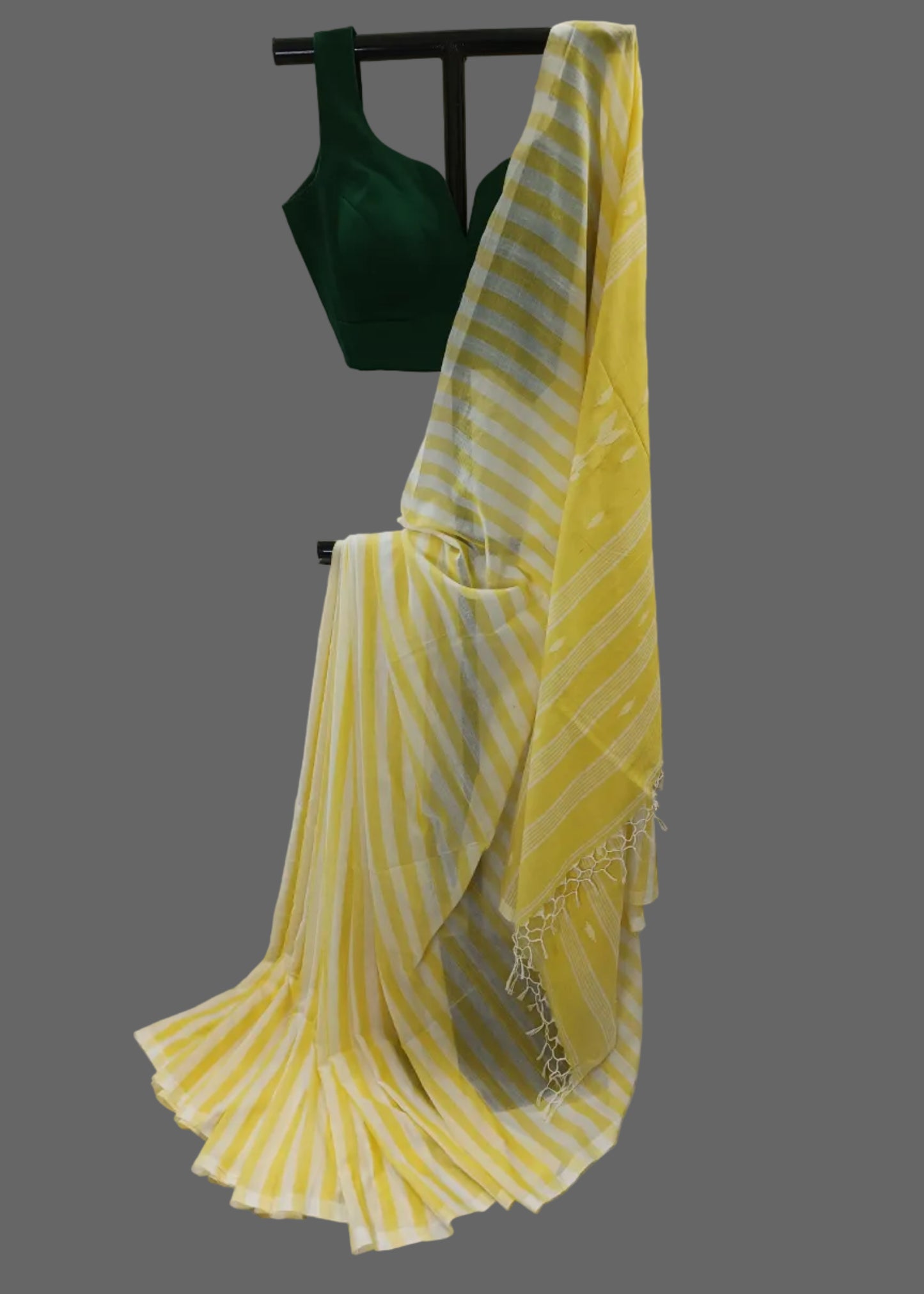 Yellow and white striped cotton saree draped on a black hanger stand, paired with a dark green sleeveless blouse, set against a draped white fabric backdrop.