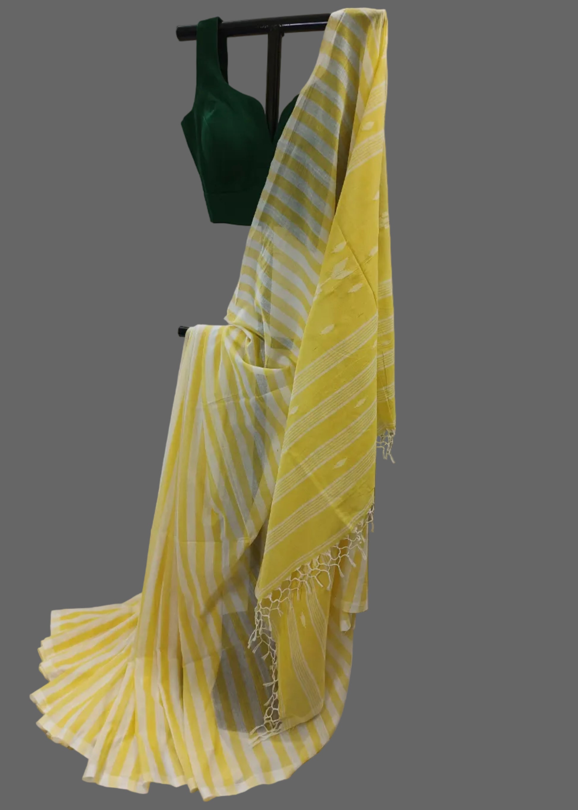 A yellow and white striped saree draped over a black clothing rack, paired with a dark green blouse, set against a draped white fabric backdrop.