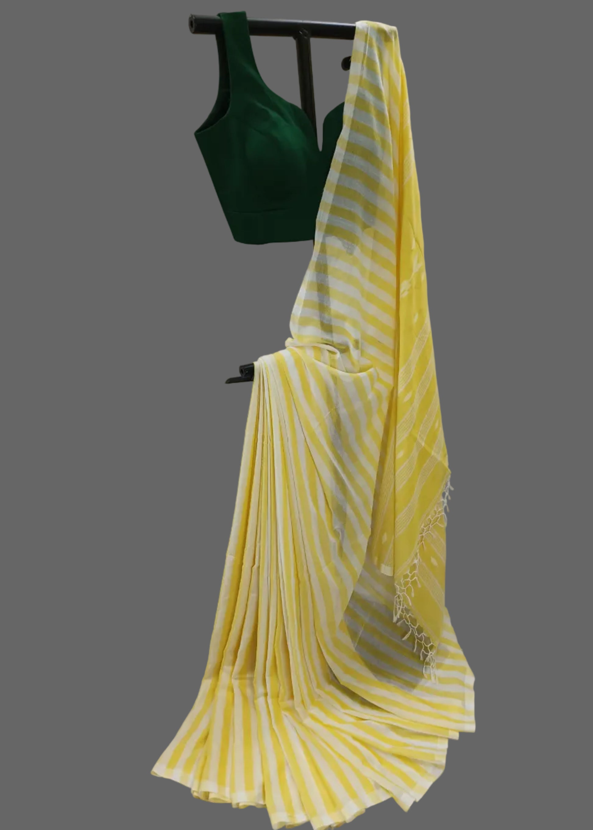 A yellow and white striped saree draped over a black hanger, paired with a dark green sleeveless blouse, set against a draped white fabric backdrop.