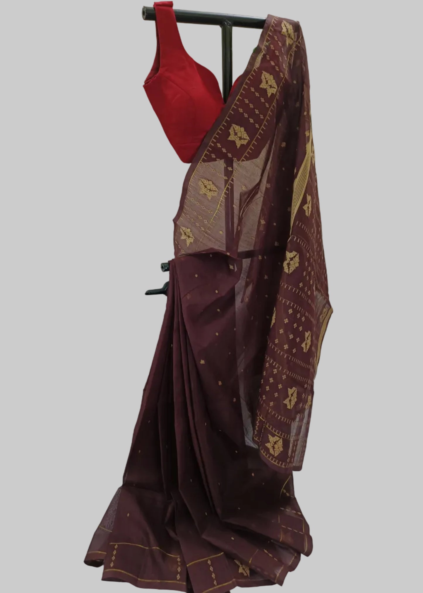 A dark maroon saree with gold patterned borders and a solid red blouse, displayed on a black clothing rack against a white draped backdrop.