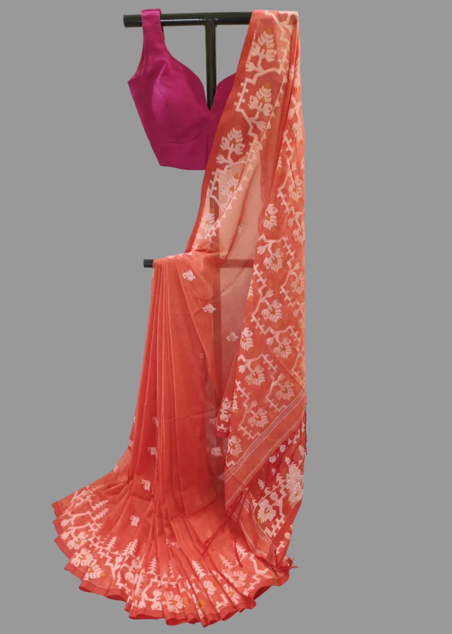 A coral orange georgette saree with white floral prints and a matching magenta pink blouse, displayed on a black clothing stand against a white fabric backdrop.