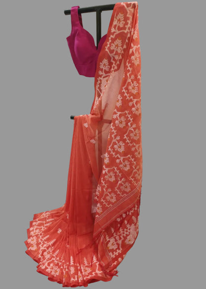 A coral orange saree with white floral bandhani patterns, draped over a black clothing rack next to a magenta pink blouse, set against a draped white fabric backdrop.