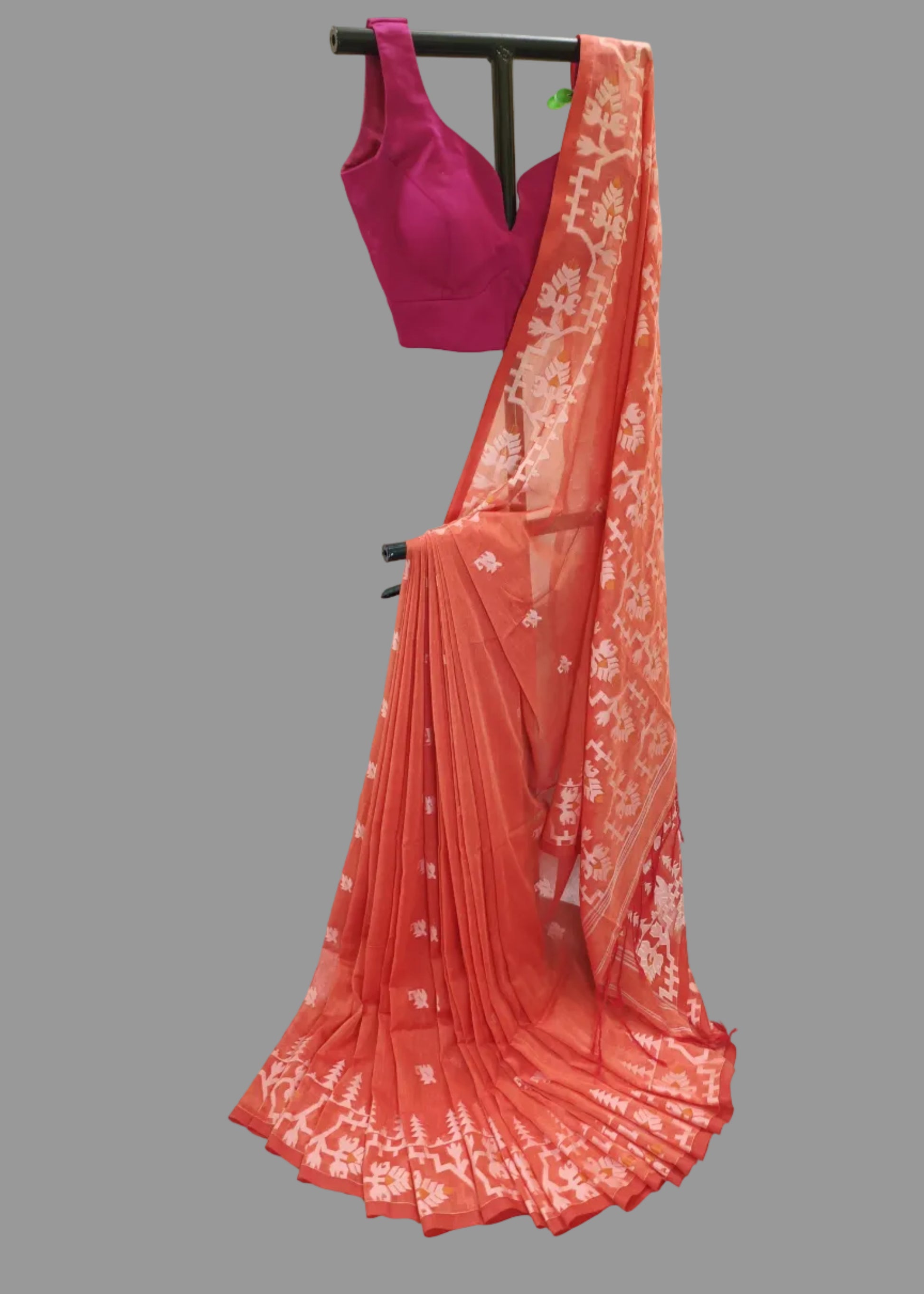A coral orange saree with white floral patterns and a matching magenta pink blouse, displayed on a black clothing rack against a white draped backdrop.